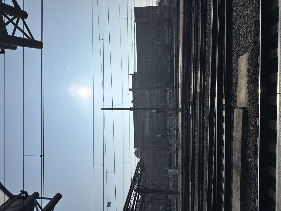 The image displays a wide view of a railway yard and platforms in Saint-Gilles - Sint-Gillis, Belgium, likely captured from a train or elevated station perspective. The scene is dominated by multiple parallel railway tracks, approximately ten sets, running horizontally across the lower two-thirds of the frame, composed of metal rails on gravel ballast and sleepers. Concrete platform sections are visible between some tracks.
In the midground, to the left, a dark, arched metal canopy structure covers a portion of a platform, adjacent to which is a graffiti-covered utility box displaying white and blue lettering. Several tall, dark utility poles are positioned between the tracks and the background building. Further back, additional platforms or concrete walls are visible, also marked with various textual graffiti.
The background is dominated by the Royal Belgian Institute of Natural Sciences building, a large, multi-story structure with a dark, gray facade and numerous windows, situated beyond the railway lines. A distinctive domed roof feature is prominent on the left side of this building. The lower sections of the building also display extensive graffiti.
The sky is clear and bright blue, featuring a prominent sun flare slightly right of center and a faint aircraft contrail in the upper right quadrant. Overhead, numerous electrical catenary lines crisscross the sky, supported by dark metal structures partially visible in the upper corners. Shadows are cast towards the right side of the frame, indicating the sun's position. No individuals are discernible within the immediate railway track or platform areas.