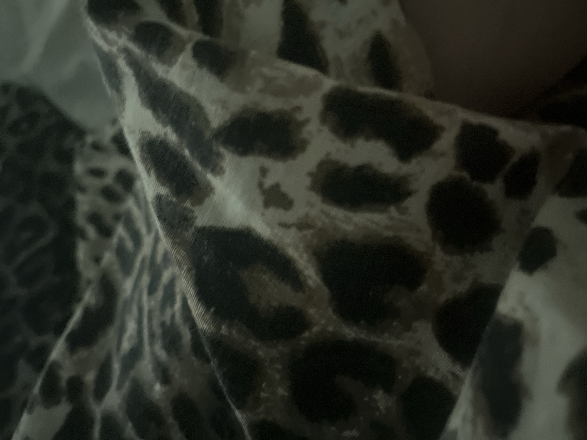The image shows a close-up view of a leopard print fabric, likely a blanket or bedsheet. The fabric is folded, creating shadows and highlights that emphasize its texture. The print consists of irregular dark brown or black spots on a lighter tan or beige background. The lighting is dim, suggesting the photograph was taken indoors in low light conditions. There are no visible people, animals, or distinct objects other than the fabric itself. The background is out of focus and appears to be a solid, dark color. The location is unknown.