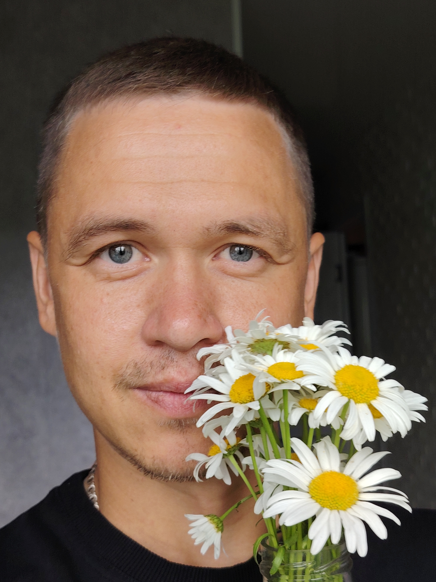 A close-up selfie features a man with light skin, short brown hair, and blue eyes. He is holding a bouquet of daisies in front of his face, with some of the flowers partially obscuring his mouth and nose. He has a light mustache and a small patch of stubble on his chin and jawline. He is wearing a black t-shirt and a silver chain necklace. The background is dark and out of focus, with a hint of a textured surface on the right side. The lighting suggests it may be daytime. The man's expression is neutral. The image is taken in Chaikovsky, Russia.