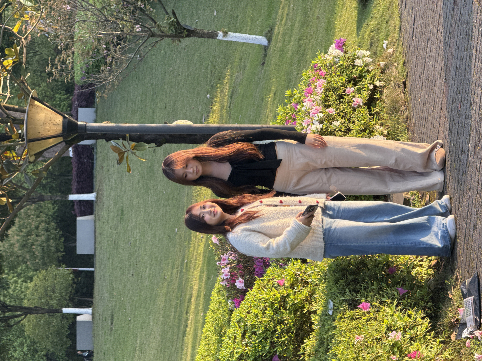 Two young women stand near a flower bed in Jiangjin, China. One woman wears a black long-sleeved shirt and beige pants. The other wears a white sweater and blue jeans. One is holding a cell phone. They are next to a paved path near a trimmed hedge with pink and white flowers. A lamp post with a decorative top stands to the left, and the background includes a green lawn and trees.