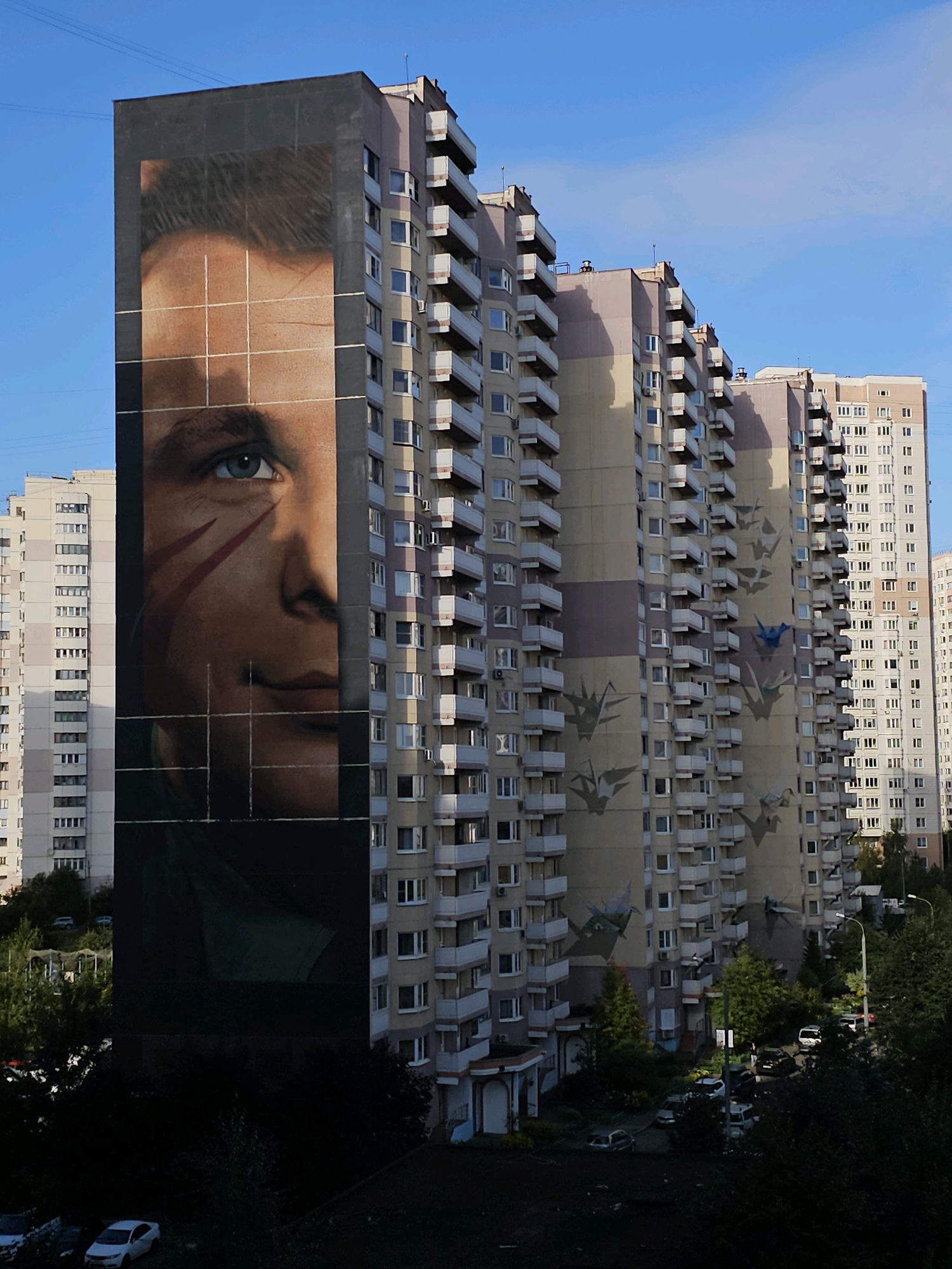 A large mural of a man's face is painted on the side of a tall, multi-story apartment building in Odintsovo, Russia. The mural covers a significant portion of the building's facade, with the man's eye and part of his face visible. The building has many windows and balconies, typical of residential architecture. Other apartment buildings and trees are visible in the background. A few cars are parked on the street in front of the buildings. The sky is clear and blue.
