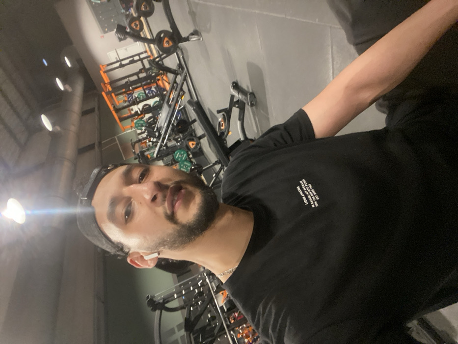 A male individual is depicted in a gym environment in Hœnheim, France. He has dark hair, a beard, and is wearing a black cap backward. A white wireless earbud is present in his left ear, and a silver chain is visible around his neck. He wears a black t-shirt with the white text "LIONS ATHLETIC IN SAVAGE / NOT AVERAGE TURN THE PAIN INTO POWER EST. 1997" printed on the chest. The man is looking directly at the camera, appearing to take a selfie. The background displays a fitness center with dark gray rubber flooring and various exercise equipment. This equipment includes weight racks with orange frames, barbells, weight plates (some green, some black with orange accents), and a flat bench. Overhead lighting illuminates the scene, with one bright light source producing a distinct lens flare near the top center. The ceiling is high, revealing exposed ductwork.