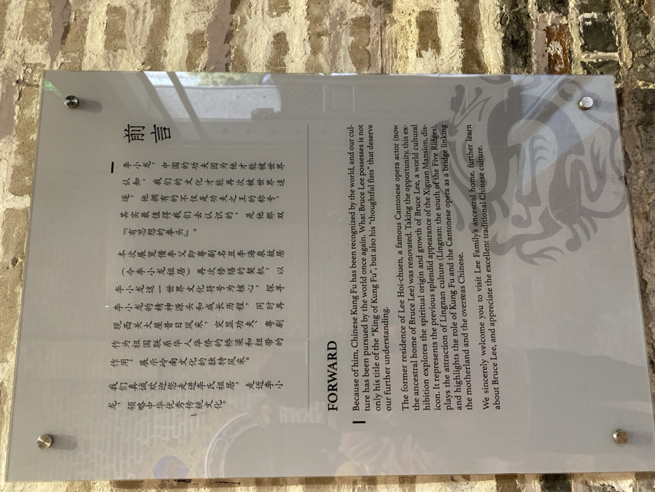 A clear acrylic or glass plaque is affixed to a rustic, light-colored brick wall, secured by four silver stand-offs. The plaque displays text in both Chinese and English, serving as a "Foreword" or "Forward" for an exhibition.
The English text, prominent on the right, explains that Bruce Lee's global recognition brought Chinese Kung Fu to the world's attention. It highlights his title as the "King of Kung Fu" and his "thoughtful fists." The text further describes the renovation of Lee Hoi-chuen's former residence (now Bruce Lee's ancestral home) and how the exhibition explores Bruce Lee's spiritual origin and growth as a world cultural icon. It also mentions showcasing the previous splendor of the Xiguan Mansion, the attraction of Lingnan culture (from the south of the Five Ridges), and the role of Kung Fu and Cantonese opera as a link between the motherland and overseas Chinese. The text concludes with a welcome to visitors to learn more about Bruce Lee and traditional Chinese culture. The Chinese text on the left conveys the same message.
A faint, stylized dragon-like watermark is visible on the right side of the plaque. The brick wall background suggests an old or historically preserved building, consistent with the description of an ancestral home and mansion. The overall setting appears to be an indoor exhibition space, likely within the Lee Family's ancestral home in Guangzhou, China.