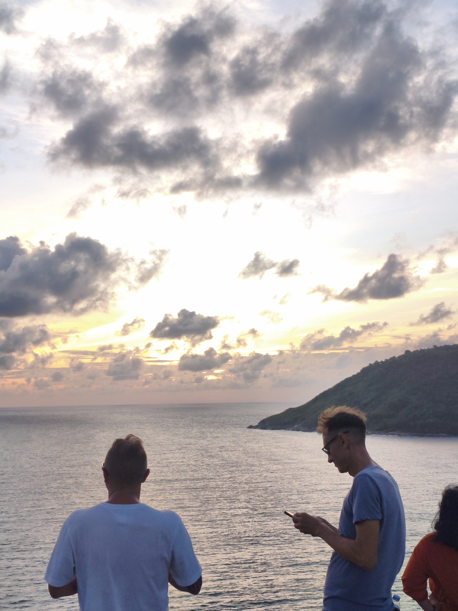 At dusk, overlooking the ocean in Rawai, Thailand, a man in a white t-shirt stands with his back to the camera, facing the water. To his right, another man with glasses is looking down at his phone. A third person, partially visible on the far right, wears an orange shirt. The sky is filled with dramatic clouds tinged with the colors of the sunset, and a lush green island rises from the sea in the distance. The water is calm, reflecting the soft light of the fading day.