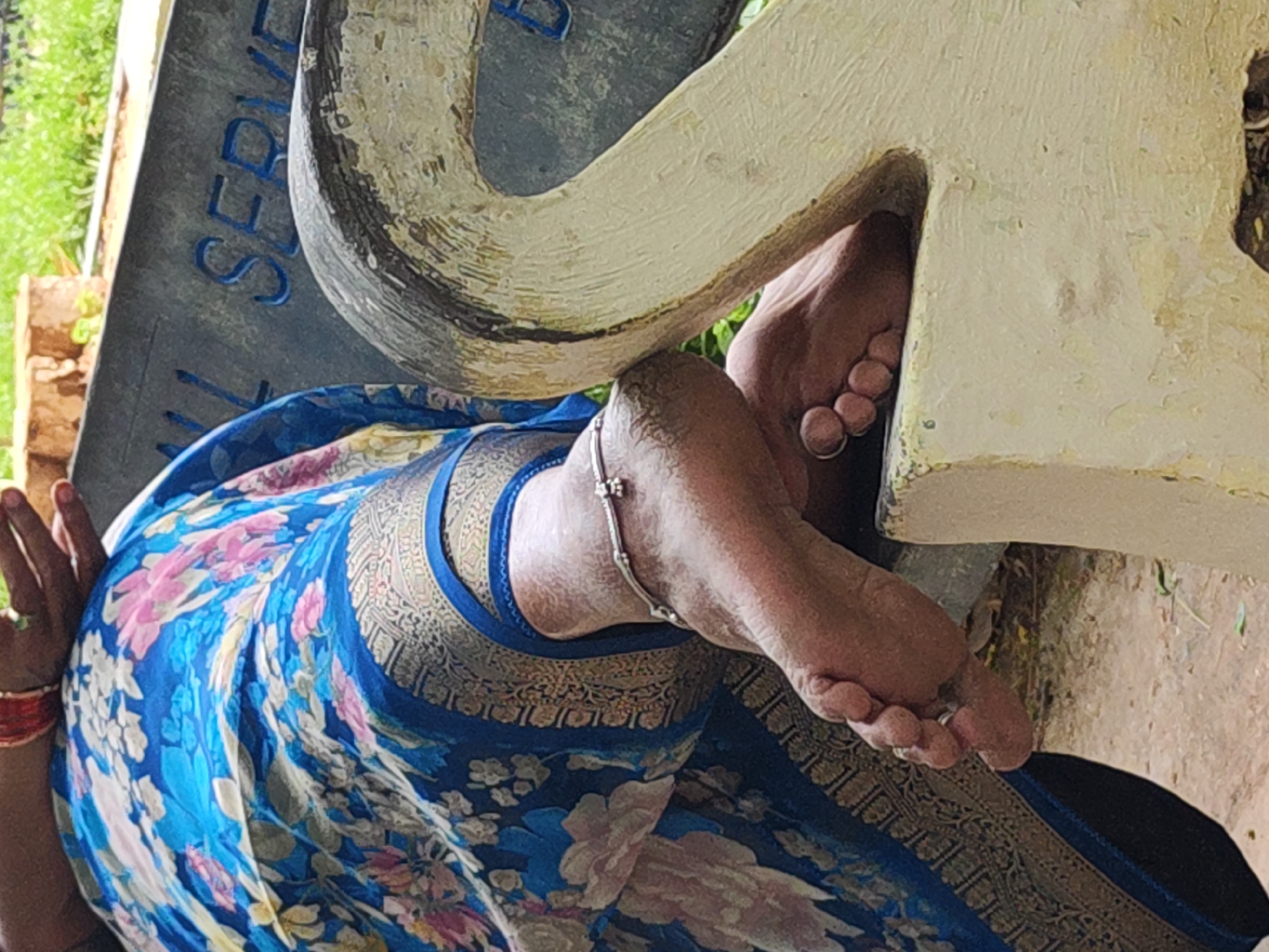 A close-up captures the feet of a person, seemingly female, adorned with a silver anklet, resting on a white painted structure, possibly part of a memorial stone. The individual is draped in a blue floral saree with a gold border. The location appears to be near a marble or granite plaque with letters partially visible. Given the context of Puttaparthi, India, this may be within the grounds of Prasanthi Nilayam or a related memorial site.