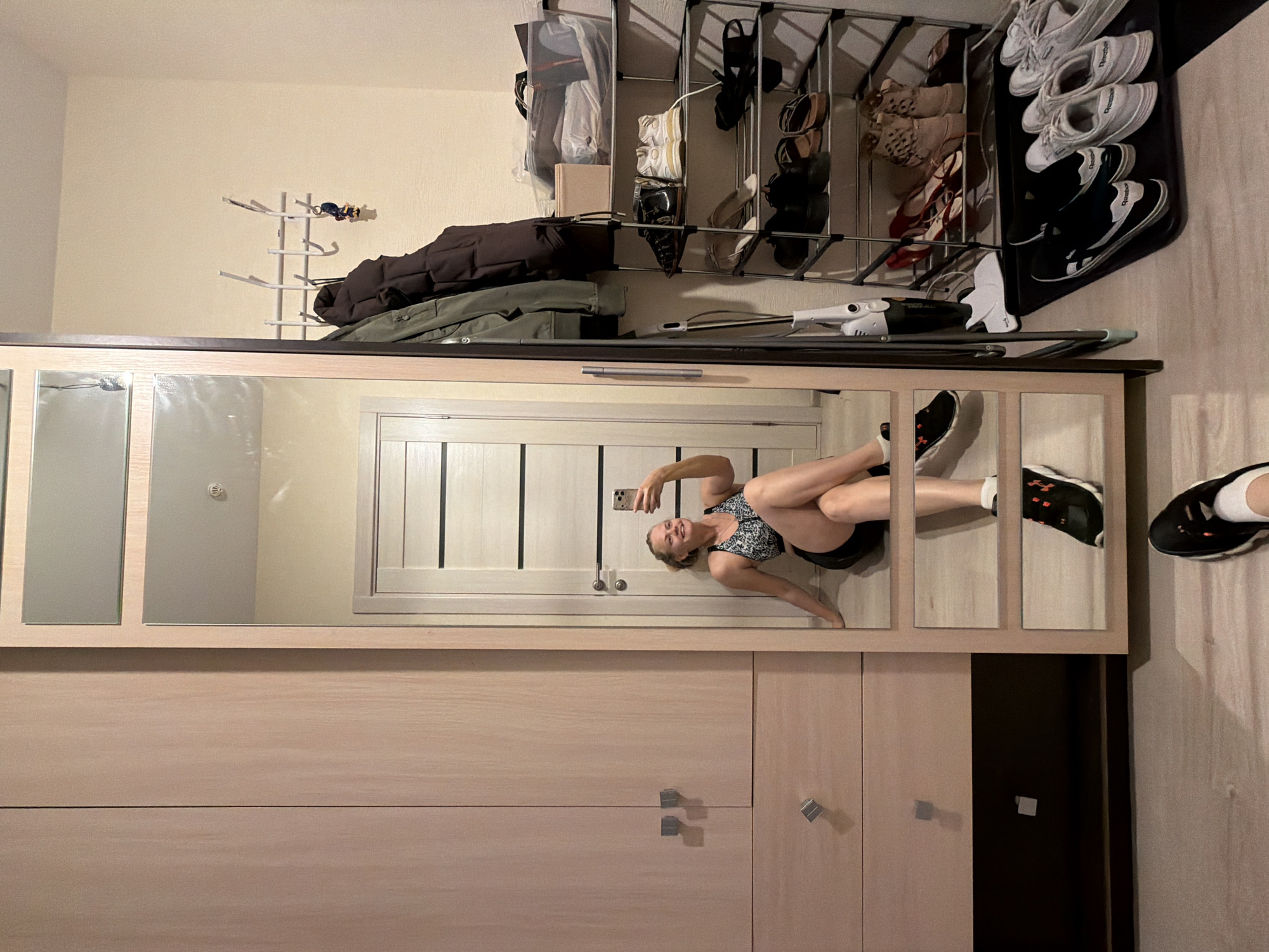 An indoor scene depicts a person reflected in a full-length mirror, surrounded by home furnishings and storage solutions.
On the left, a tall, light-toned wooden wardrobe or closet features two upper doors and two lower drawers, each fitted with square, metallic handles. The base of the wardrobe is dark brown.
In the center, a full-length mirror, framed in light wood and segmented by horizontal dividers, reflects a female seated on the floor. The reflected person has light-colored hair pulled back, is smiling, and is holding a smartphone in her right hand, appearing to take a selfie. She is wearing a black and white patterned sports bra and black shorts, with her legs crossed at the knees. Behind her in the reflection is a light-colored wooden door with horizontal dark stripes and a door handle. A ceiling-mounted smoke detector is visible in the reflection above her head. On the actual floor in front of the mirror, a person's foot, wearing a black sneaker with a white sole and orange accents, is visible, suggesting the perspective of the actual photographer.
To the right of the mirror, against a light-colored wall, a white metal coat rack with multiple hooks holds an olive green jacket and a dark brown puffer jacket. A keychain with a small figure hangs from one hook. Below the coat rack, a multi-tiered black metal shoe rack is filled with various footwear. The top tier contains a clear plastic bin with contents and a small light-colored box. Subsequent tiers display a variety of shoes including dark sneakers, white sneakers, light-colored sandals, dark sandals, dark shoes, brown sandals, red flats, and patterned boots. Leaning against the wall between the mirror and the shoe rack is a white stick vacuum cleaner. On the light-toned wooden floor in front of the shoe rack, a black shoe tray holds several pairs of sneakers, including black and white Reebok, white Reebok, and light grey/white pairs. The floor throughout the visible area is consistent light-toned wood or laminate.