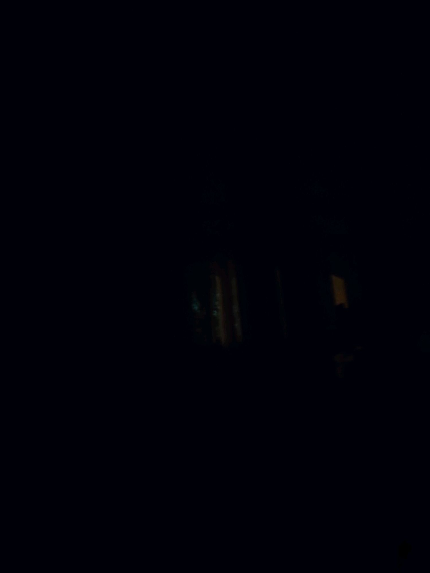 The image is a dark, blurry shot of what appears to be a building or structure at night. Several illuminated windows or openings are visible, with one showing a vertical striped pattern that is brightly lit with orange and green hues. To the right, a rectangular orange light source is visible. The scene suggests an urban environment, possibly residential or commercial. Without further context or clearer details, it is impossible to determine the specific location in Severodvinsk, Russia, or to identify any people, actions, or specific objects beyond the illuminated openings in the structure. The overall impression is one of low light conditions and a focus on the contrast between the dark surroundings and the artificial lights.