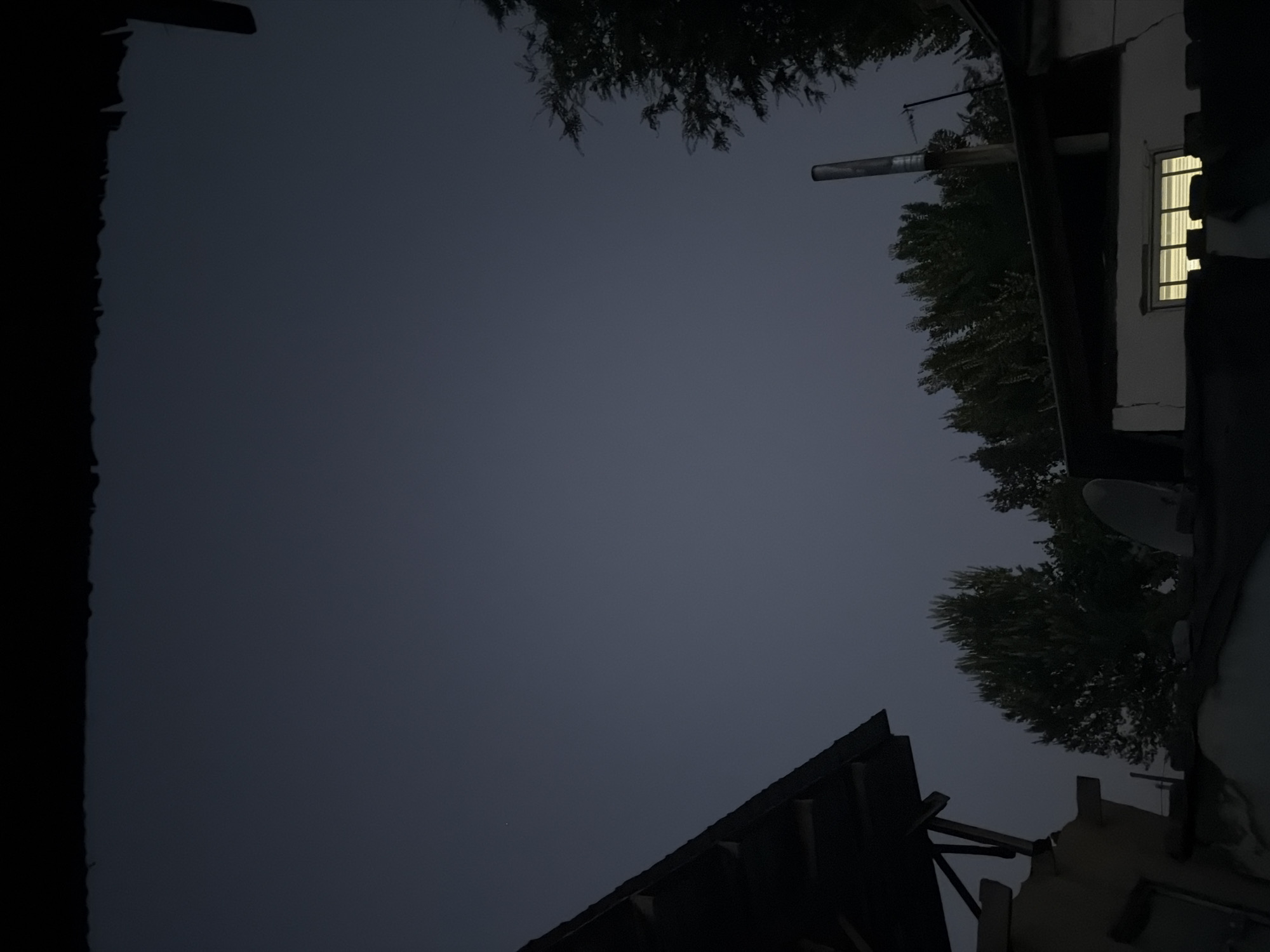 The image captures a dimly lit outdoor scene, possibly during twilight or early night, in Belovodskoe, Kyrgyzstan. The sky is a dark, overcast grey. In the foreground, the roof of a building juts into the frame from the left, appearing dark and shadowed, with visible wooden support beams underneath. To the right, a section of another building is visible, featuring a window with illuminated blinds, suggesting interior light. A metal chimney pipe stands vertically on the roof of this building. A cluster of trees with dark foliage lines the horizon behind the buildings, their shapes softened by the low light. A small, faint white object, possibly a distant star or satellite, is visible in the upper left portion of the sky. The overall atmosphere is quiet and still.
