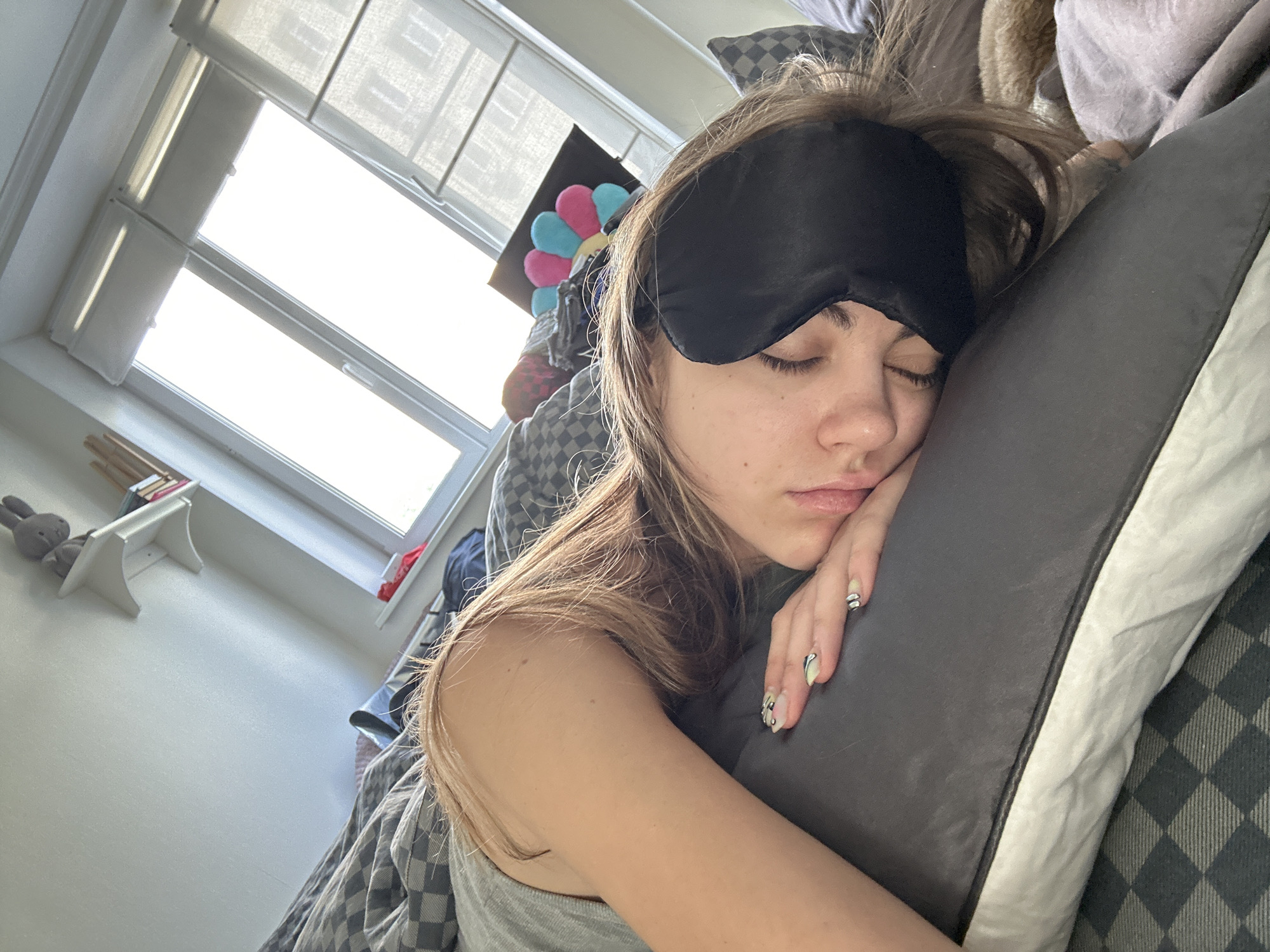 A young Caucasian female with long, light brown hair is lying asleep on a pillow in a bedroom. She is wearing a grey tank top and a black sleep mask covering her eyes. Her right arm is bent and resting on the pillow, and her fingernails are painted with a light green and white design. She is wearing a silver ring on her left index finger. The room appears to be a bedroom with a window behind her, letting in bright sunlight. There are patterned blankets on the bed, and a shelf with a stuffed rabbit and books is visible on the wall. A colorful flower-shaped cushion is also present in the background. The overall scene depicts someone sleeping in a domestic setting.