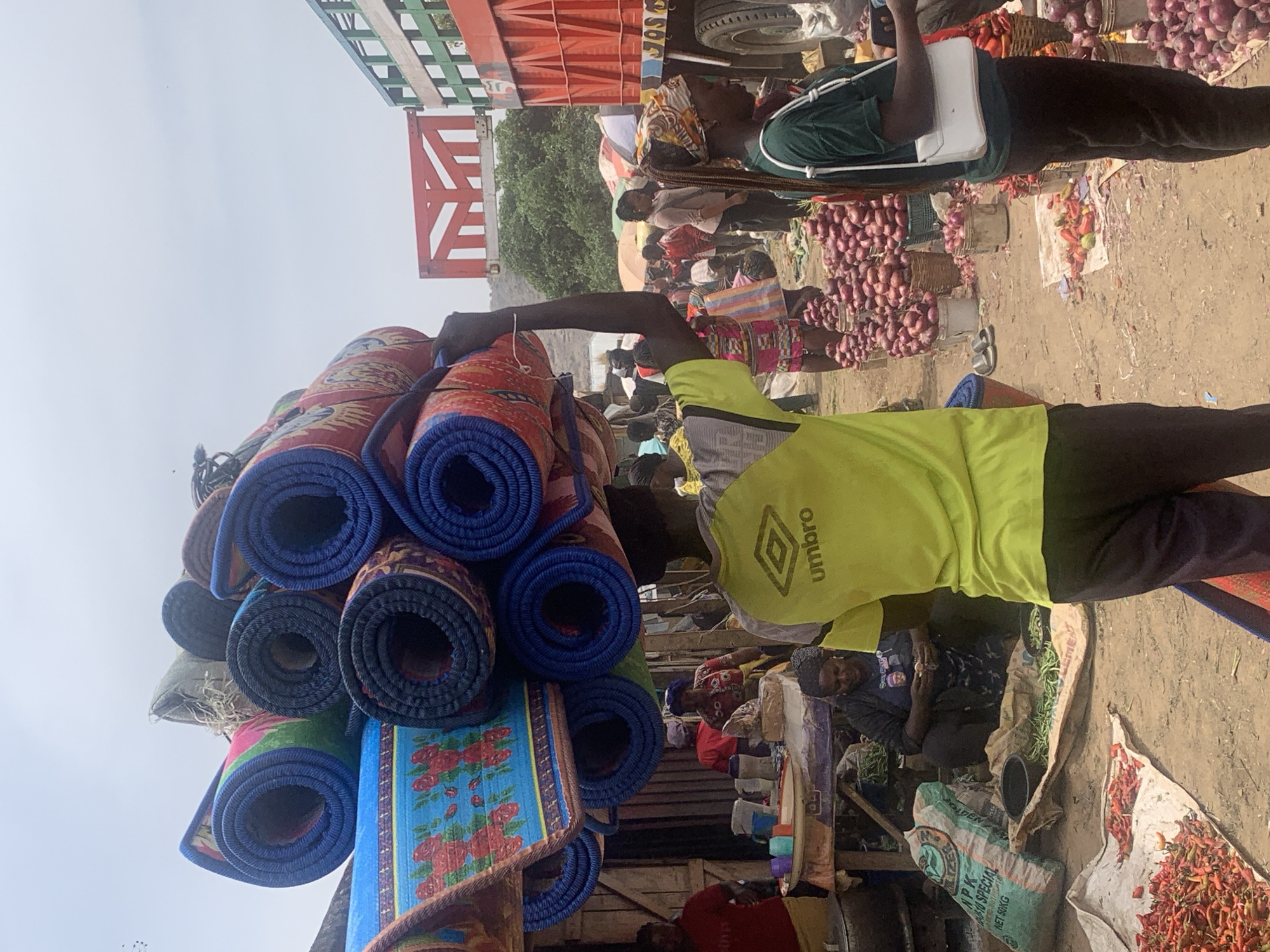 The image depicts a bustling marketplace in Jos, Nigeria. A man wearing a bright yellow Umbro t-shirt is carrying a stack of rolled-up mats. Another person, partially visible, wears a green shirt and a white crossbody bag, stands near piles of onions. The ground is dusty, and various goods are displayed on the ground and on tables. A red truck is partially visible in the background. Other individuals are present in the crowded market scene.