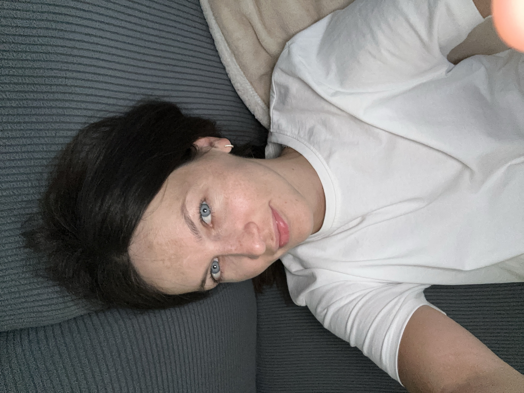 A woman with dark hair and blue eyes is lying on a textured grey couch. She is wearing a white t-shirt and a small hoop earring in her left ear. Her head is tilted back, and she is looking towards the camera with a slight smile. Her skin is fair with some freckles visible on her nose and cheeks. A beige blanket is partially visible behind her to the right. The scene appears to be indoors. This image was captured in Donji Pobori, Montenegro.