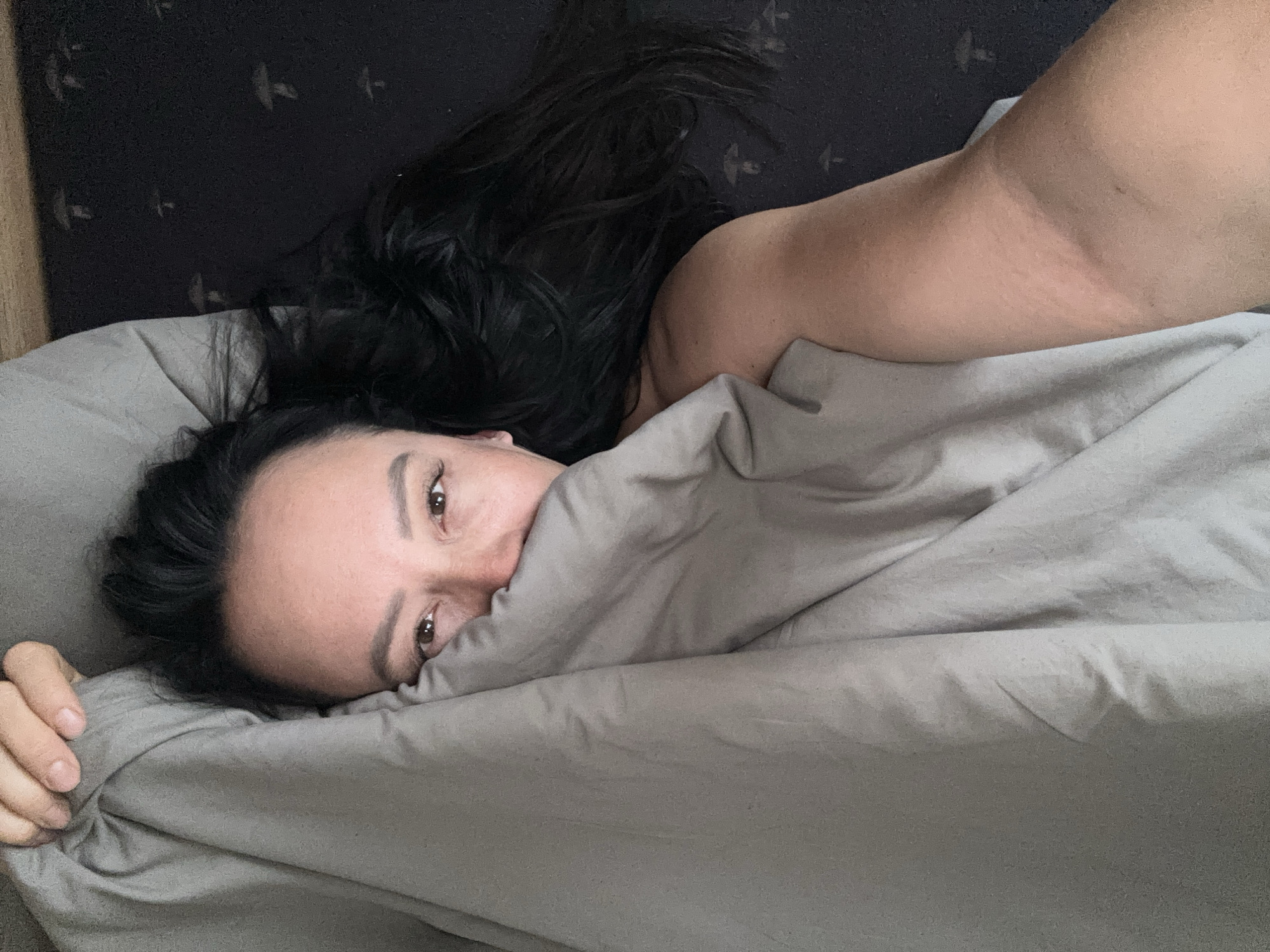 A woman with long, dark hair lies in a bed covered with gray sheets, partially obscuring her face. She is looking directly at the camera. The background features a dark, patterned wall. The location is Chaikovsky, Russia.