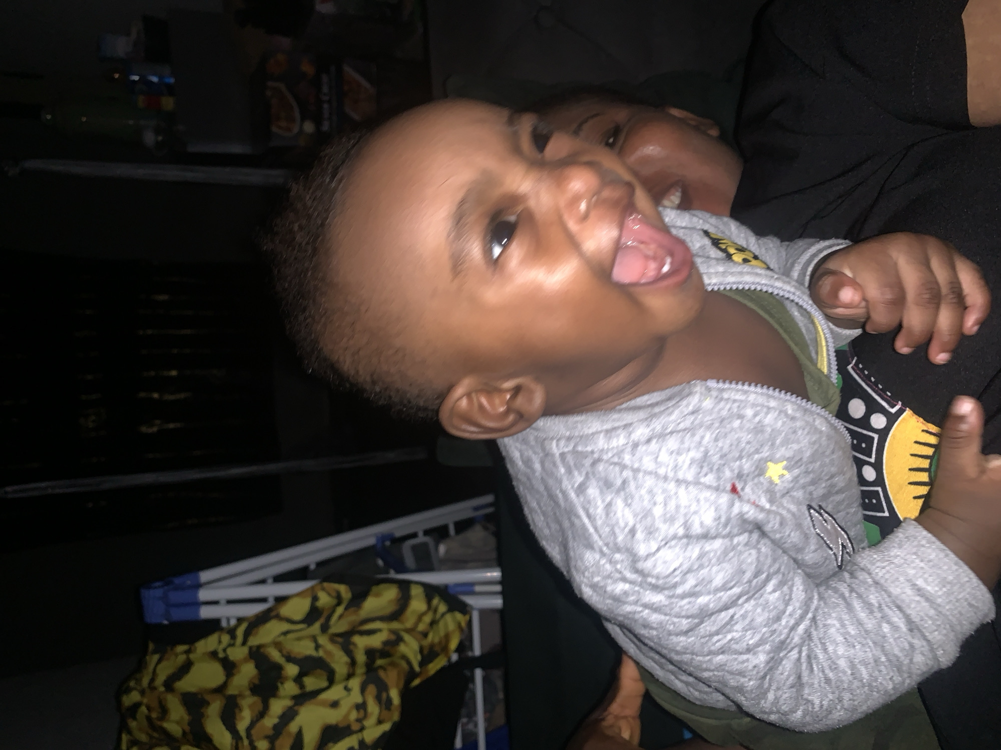 In the dimly lit interior of a residence in Barakin Akawo, Nigeria, a close-up shot captures a young child with their mouth wide open, sticking out their tongue. The child, wearing a grey quilted jacket over a green shirt and dark pants, is positioned in the foreground, their face contorted in an expression of playfulness. Behind the child, a second individual, likely an adult, is partially visible, their mouth also open in a smile or laugh.
The background is dark and indistinct, but a stepladder and a yellow and black patterned fabric are visible to the left. To the right, shelves with various items, possibly food products and bottles, can be seen. The overall impression is one of a casual, indoor setting, likely a living room or bedroom, where a moment of joyful interaction between two individuals is being captured.