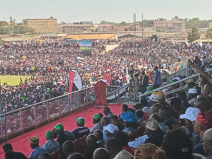 This image depicts a large outdoor public gathering or rally taking place in a stadium or arena under clear, sunny daytime conditions. The setting is consistent with an event in Siaya, Kenya.
The scene is dominated by an immense crowd of people, filling the stadium seats, standing on the field, and extending far into the background. In the immediate foreground, a section of stadium seating is visible, occupied by several individuals mostly with their backs to the viewer, facing a red-carpeted stage or platform. Among these spectators, one person wears a green cap with the text "GENOWA" clearly visible on the back, while another has "CID" partially legible on a dark cap or headband.
On the red-carpeted platform, a central figure, dressed in a red robe or garment, stands as if addressing the crowd. Nearby, other individuals, including security personnel or event staff, are present, along with professional cameras on tripods. Two prominent vertical banners are positioned near the stage; one is red with a white logo and indistinct text (possibly "B O A"), and the other is white with faint dark text (potentially "AKU"). In the distance, above the densely packed crowd, a large video screen displays content.
Beyond the stadium grounds, a cityscape or urban sprawl with various buildings and structures lines the horizon. The overall impression is that of a significant public event, likely a political rally or large community gathering, characterized by a highly engaged and attentive audience.