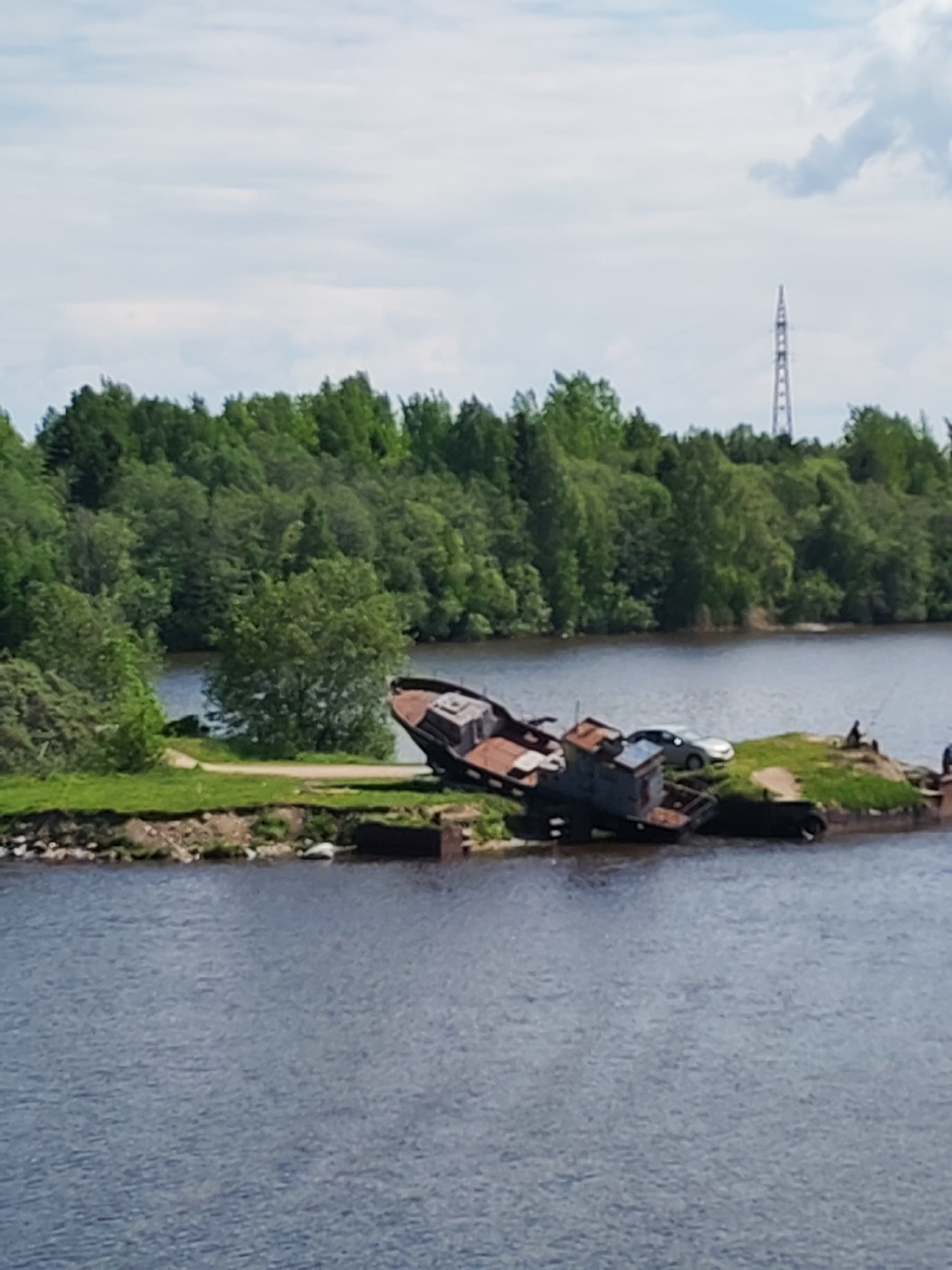 The image shows a partially submerged, decaying ship on a small, grassy island in a body of water, likely a river or lake. The ship appears rusted and dilapidated. On the island, a silver car and what appears to be a black vehicle are also visible. Two individuals are present on the island, one seemingly fishing. In the background is a dense forest and a tall electrical tower. The location is specified as Ivanov Bor, Russia.
