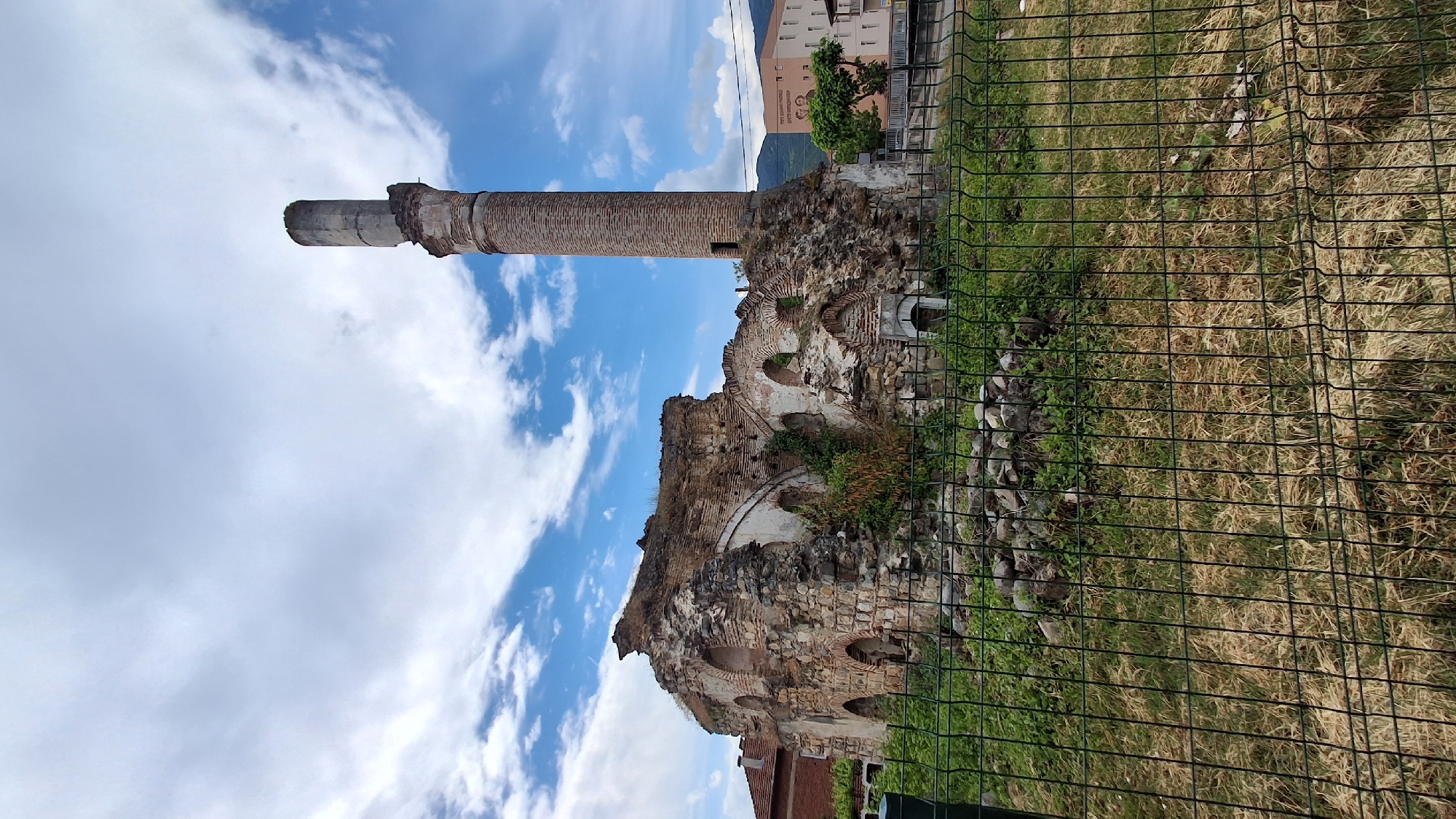 This image, set in Gotse Delchev, Bulgaria, presents a historical ruin contrasting with modern elements under a partly cloudy sky. The photograph is taken from an eye-level perspective, looking across an overgrown area.
In the immediate foreground, a dark green metal wire fence extends across the bottom and right side of the frame, partially obscuring a patch of dry grass and some scattered stones.
The central focus is a tall, slender brick structure resembling a minaret, standing prominently on the left side of the frame. Its upper section appears damaged or incomplete, with eroded stonework visible. Adjacent to and below this structure are the extensive ruins of an old building, constructed from a mix of stone and brick. These ruins feature several arched openings and crumbling walls, with patches of green vegetation and small bushes growing within the crevices and on top of the decaying masonry.
In the background, behind the ruins and minaret, modern, multi-story buildings are visible, indicating an urban or semi-urban setting. On one of these buildings, faint Cyrillic text can be discerned, reading "СУ ЗА СПОРТНО УЧИЛИЩЕ ЗА ПРАКТИЧЕСКИ СПОРТОВЕ" (SU for Sports School for Practical Sports). A green tree stands near these buildings. The sky above is a bright blue, dotted with scattered white, fluffy clouds, suggesting a clear and bright daytime.
No people or explicit activities are taking place; the scene captures a static architectural landscape that bridges historical decay with contemporary development.