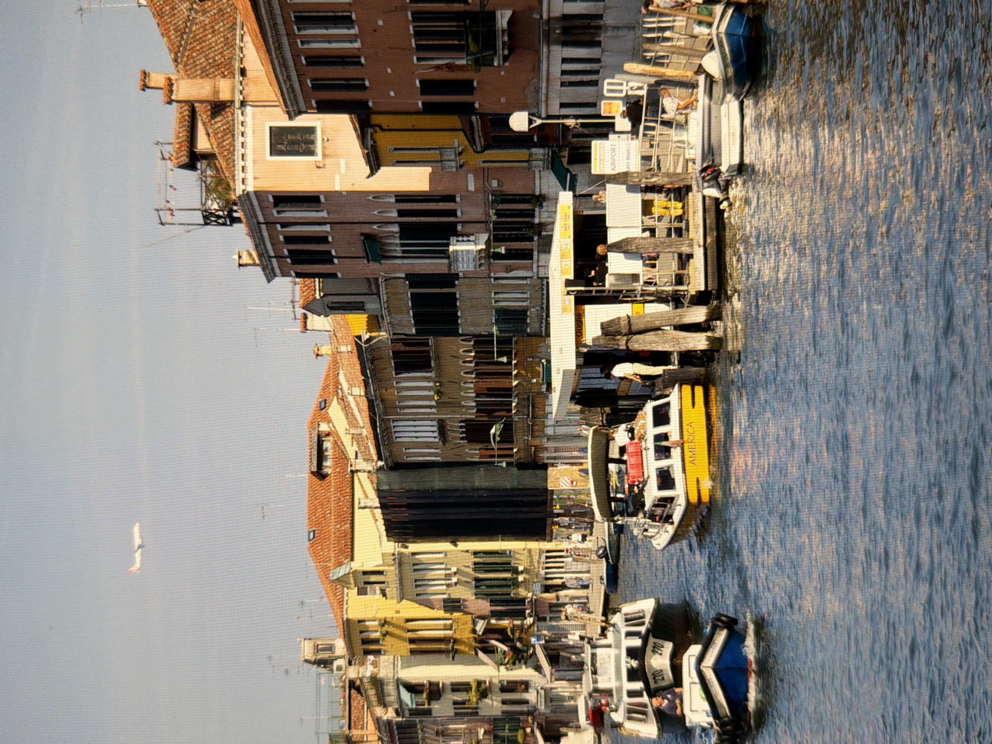 The image depicts a wide canal in Venice, Italy, with boats navigating the waterway and historic buildings lining the sides. In the foreground, several boats are visible: a white and grey boat with the numbers "270 270" on its stern is moving away from the viewer on the left. To its right, a smaller blue and white boat is cutting through the water. Further to the right, a yellow and white boat labeled "AMERICA" is docked. Behind the boats, a modern, white structure with a roof is situated at the water's edge, serving as a water bus or ferry stop, indicated by a sign that reads "AIRPORT SAN MARCO." People are seen on and around the boats and the pier. The background is filled with colorful, multi-story buildings with red-tiled roofs, characteristic of Venetian architecture. An airplane is flying in the clear blue sky overhead. The water is calm and reflects the sunlight and the surrounding structures. The scene captures a busy moment in a canal in Venice, Italy, likely during the daytime.