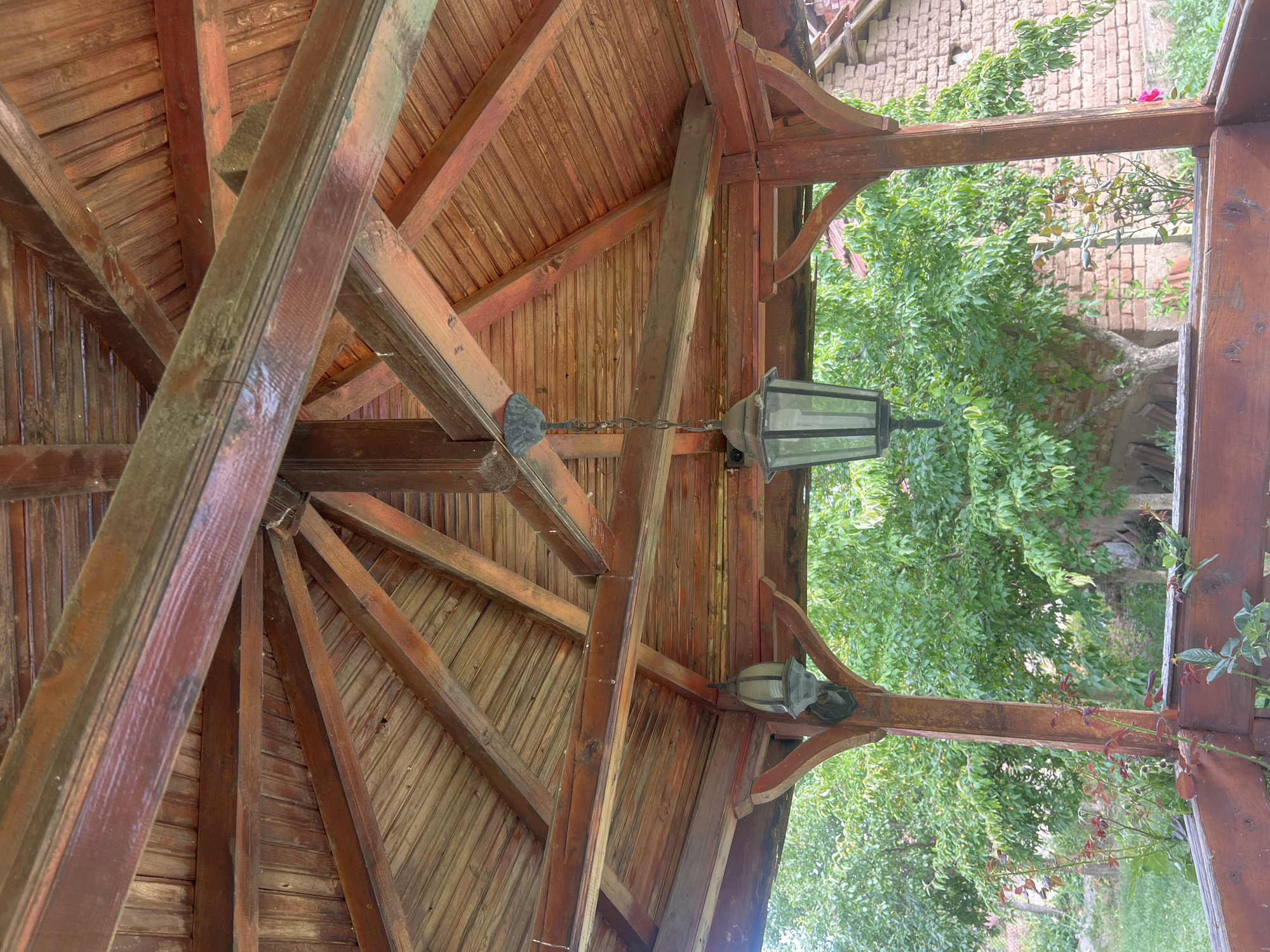 The image shows the interior of a wooden gazebo. The gazebo has a gabled roof made of wooden planks, supported by several wooden beams and struts. Two old-fashioned lanterns hang from the ceiling of the gazebo. One lantern is directly in the center of the image, hanging from a chain, while the other is to the left. The wooden structure appears weathered and aged. Through the open sides of the gazebo, lush green trees and foliage are visible, along with a brick building in the background. The scene depicts an outdoor setting, possibly a garden or a rural area.