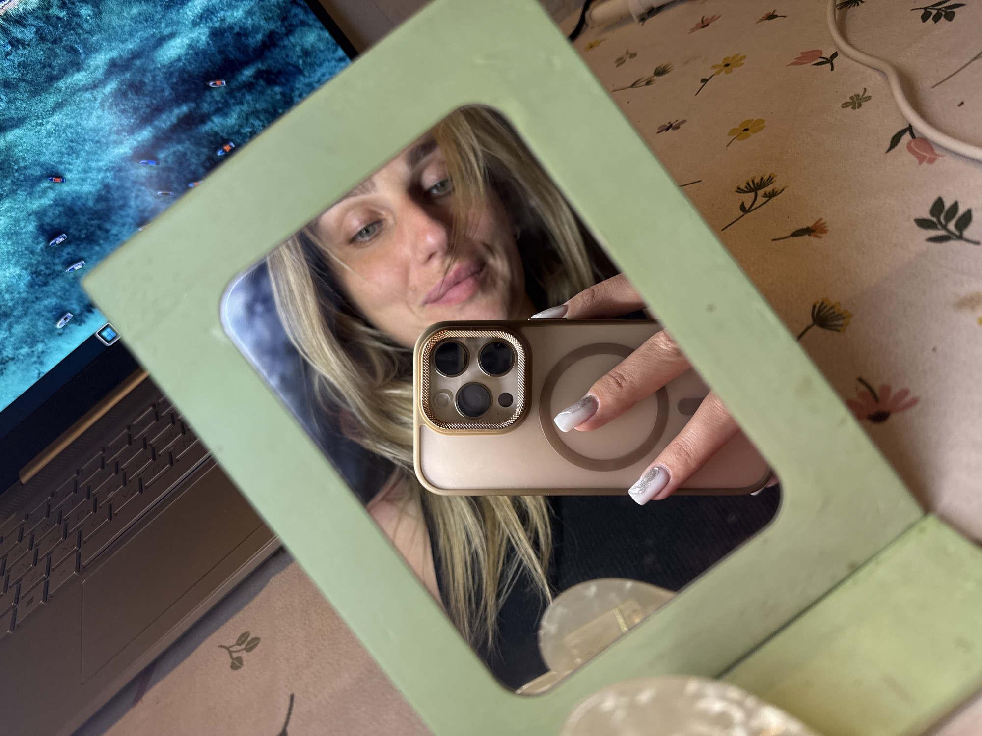 A selfie is taken in a light green frame, reflecting a woman with blonde hair and blue eyes. She is holding a gold-colored smartphone in her right hand, with her nails painted white and adorned with silver accents. Behind her, a laptop is visible, along with a screen displaying a vibrant turquoise ocean with several small boats. The frame of the mirror rests on a surface covered with a light-colored cloth featuring a floral pattern with small yellow, pink, and orange flowers and green leaves. The overall scene appears to be indoors, possibly in a personal living space.