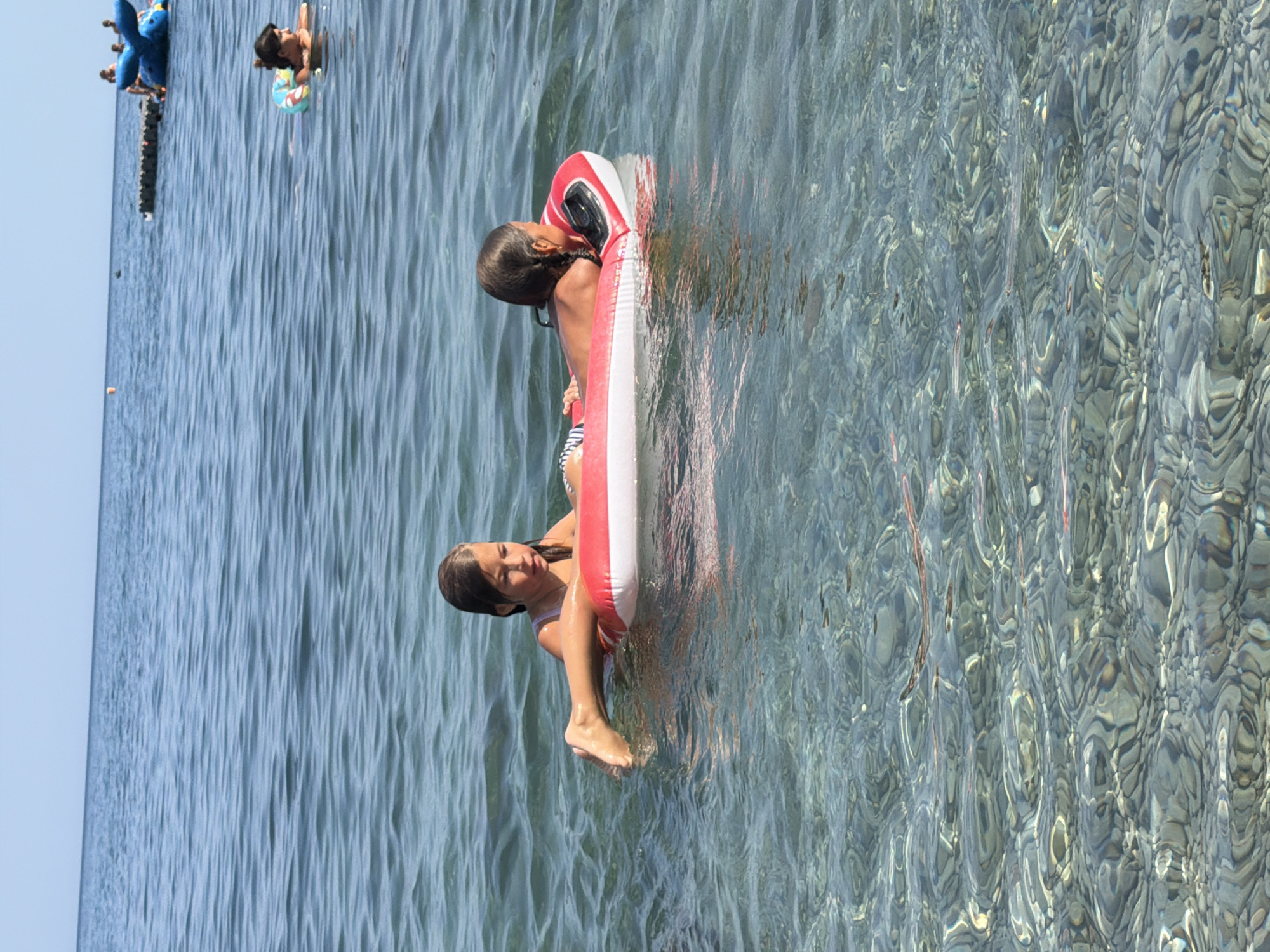 Two young girls are on a red and white inflatable raft in clear, shallow water. The girl on the left is leaning back, with her legs extended, while the girl on the right is lying on her stomach, facing away from the camera. The water is transparent, revealing smooth, rounded stones on the seabed below. In the background, the sea extends to the horizon under a clear blue sky, with a few distant figures and inflatable structures visible. The scene appears to be a beach in Partenit, Ukraine, on a sunny day.