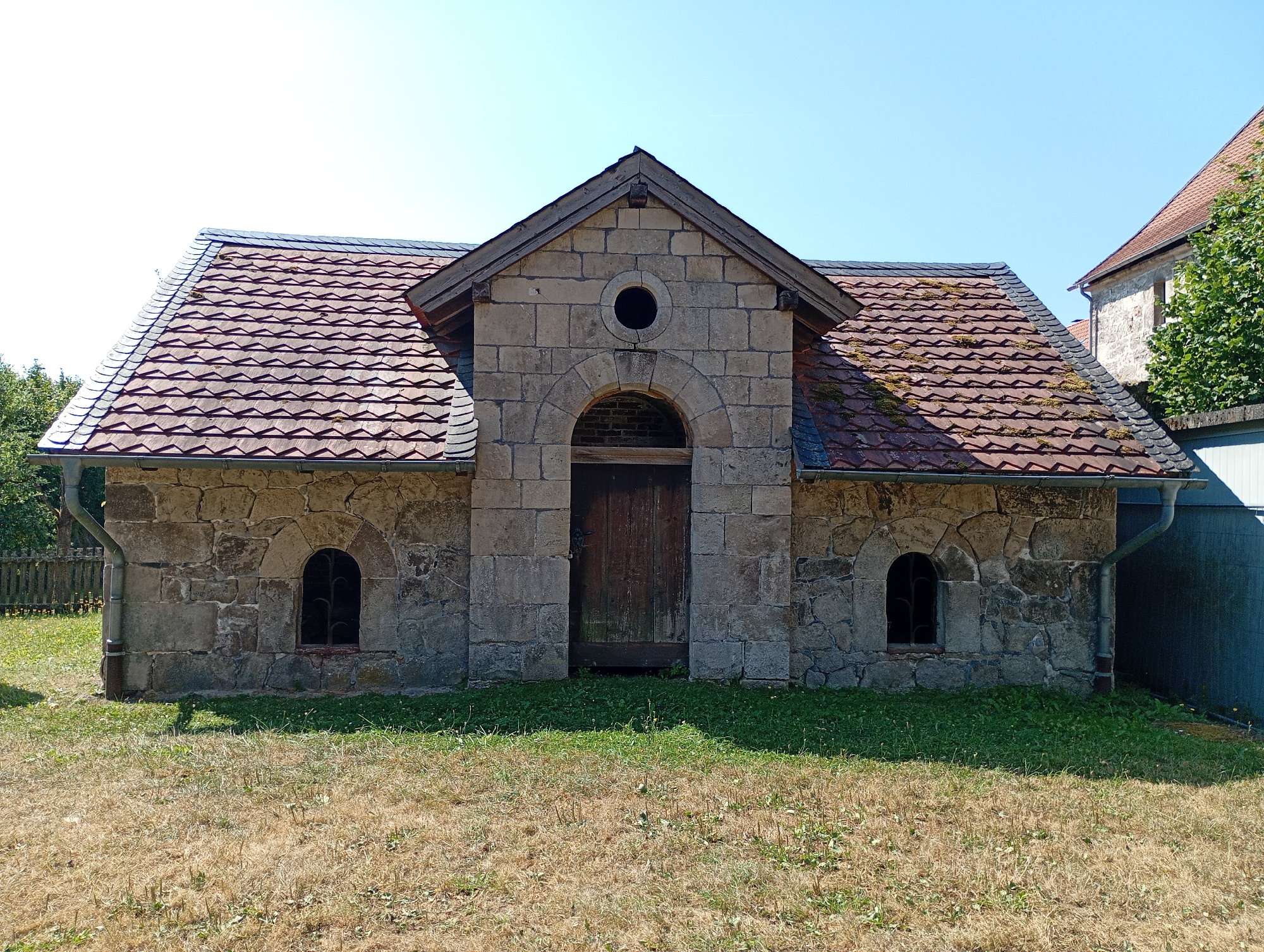 This is a full-shot, eye-level photograph of a small, stone building situated in a grassy area. The building has a gabled roof covered with reddish-brown tiles that appear to be moss-covered in places. The facade is constructed from rough-hewn grey stones. A large, arched wooden door is centrally located, flanked by two small, arched windows with metal grates. Above the arched doorway, there is a circular opening. The building is set against a clear blue sky. The foreground consists of dry, yellowed grass, transitioning to greener grass closer to the building. To the left of the building, a wooden fence is visible. To the right, a portion of another building and some green foliage are present. The building is likely a historical structure, possibly a shed or outbuilding, located in the vicinity of the Walkenried Abbey in Walkenried, Germany. No people or animals are present in the image.