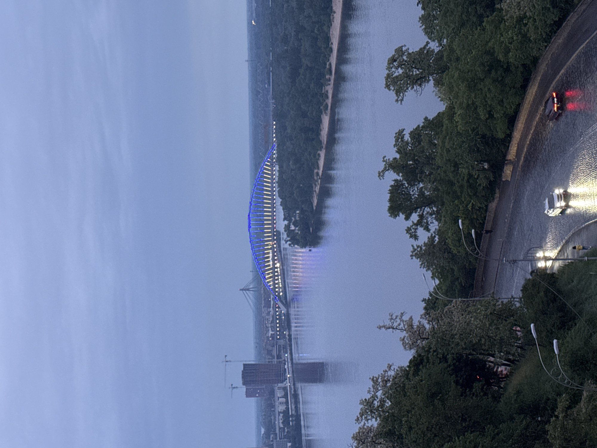 The image presents an elevated, wide-angle view of a city scene at dusk or early night, likely overlooking the Dnieper River in Kyiv, Ukraine. A prominent, architecturally distinctive bridge, possibly the Kyiv Pedestrian Bridge, spans across a wide body of water. The bridge is brightly illuminated with a combination of blue and white/yellow lights, creating reflections on the water's surface.
On the left side of the river, the distant bank is densely forested, with a narrow sandy strip visible along the waterline. Further into the distance, beyond the main illuminated bridge, other bridges, urban buildings, and construction cranes can be faintly discerned against the muted sky.
On the right side of the frame, a curved, wet road or highway runs parallel to the river, bordered by lush green trees. Two vehicles are visible on this road: a white car with its headlights on, and a dark car with red taillights illuminated, both reflecting on the damp asphalt. Several streetlights are also active along this road. A solitary figure appears to be walking near the white car.
The sky is a uniform, pale blue-grey, indicating an overcast evening or recent rainfall, contributing to the wet appearance of the road. The overall ambiance is serene, characterized by the soft glow of artificial lights against the twilight gloom. No visible text is present in the image.