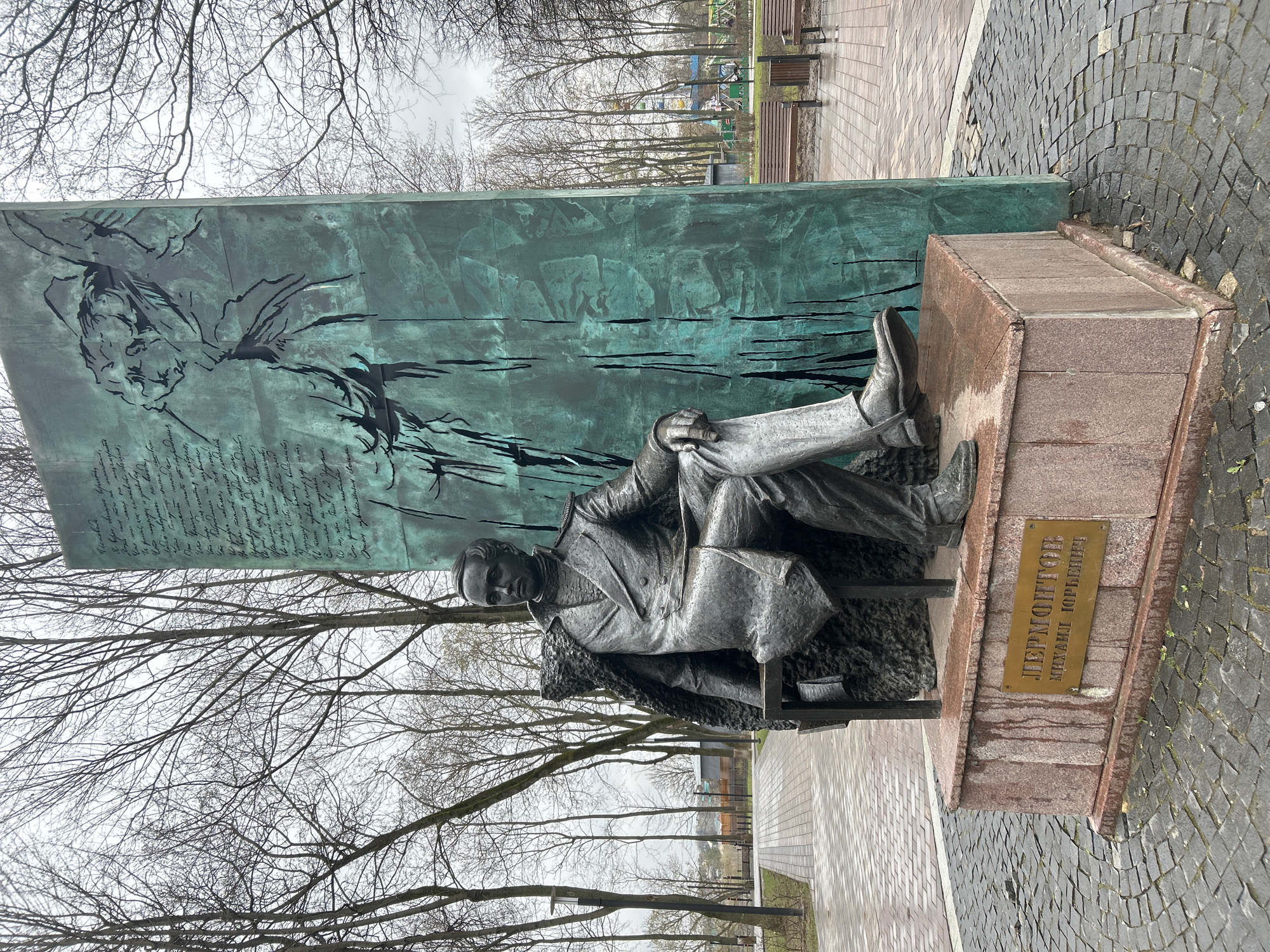 The image displays an outdoor monument to Mikhail Lermontov, a renowned Russian writer, located in Podolsk, Russia. The central subject is a dark-colored statue of Lermontov, depicted seated in a contemplative pose with one leg crossed, wearing a long coat with a fur collar. He appears to be holding papers or a book in his hands.
Behind the statue stands a tall, rectangular panel, heavily patinated with a distinctive verdigris color, suggesting aged bronze or copper. The upper left of this panel features an etched or embossed artistic design, including a profile of a face and swirling, abstract lines. Below this design, handwritten script is etched into the metal, likely excerpts from his literary works.
The statue rests on a low, reddish-brown granite or stone pedestal. A brass plaque on the pedestal clearly identifies the subject in Cyrillic as "ЛЕРМОНТОВ / МИХАИЛ ЮРЬЕВИЧ" (Lermontov / Mikhail Yuryevich).
The scene is set outdoors in what appears to be a park or public square. The sky is overcast and grey, creating diffused lighting. Numerous bare deciduous trees surround the monument, indicating late autumn, winter, or early spring. The ground is paved with a mixture of dark cobblestones and lighter, regular paving stones. In the background, visible through the trees, are park benches and other elements of a recreational area. No specific event is taking place; the image captures the monument itself in its environment.
