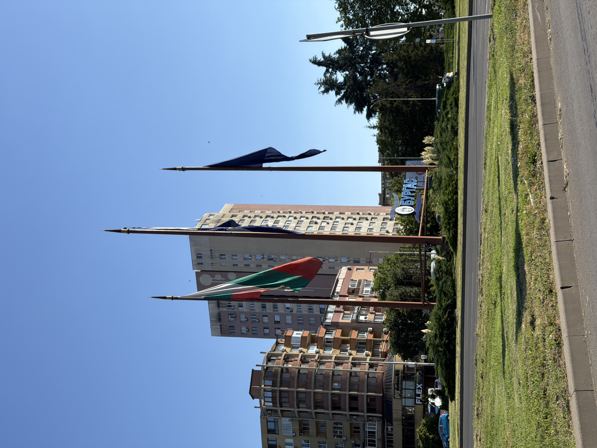 This is a vertical, eye-level shot of a city street scene in Burgas, Bulgaria, on a bright, clear day. The foreground features a paved road and a well-maintained grassy median strip bordered by a concrete curb. Beyond the median, several multi-story buildings dominate the midground and background.
A prominent feature is a large sign with blue lettering that reads "БУРГАС" (Burgas). Two flagpoles are visible, each displaying a flag. The lower flag is a tricolor of green, white, and red, horizontally oriented. The upper flag appears to be dark blue. Behind the flags, buildings of varying architectural styles and heights rise against the clear blue sky, some with visible windows and air conditioning units. Trees and more greenery are interspersed throughout the scene, suggesting a park-like or landscaped urban environment.
The lighting suggests it is daytime, likely morning or afternoon, given the bright sunlight and clear skies. The scene is calm and orderly, conveying a sense of urban life. No people are clearly visible, but the presence of vehicles further down the street hints at ongoing activity.