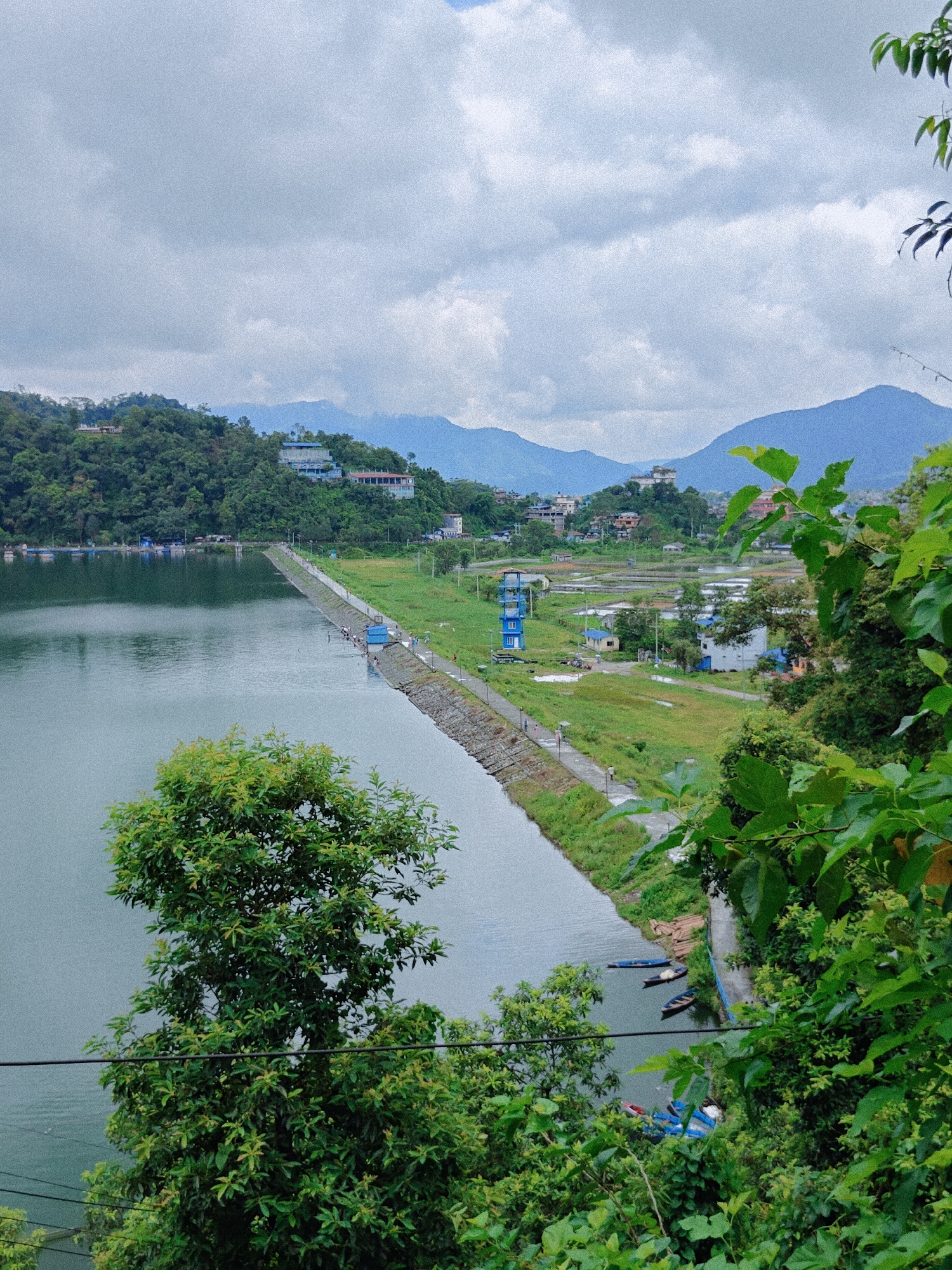 The image depicts a scenic view of Phewa Lake in Pokhara, Nepal, on a cloudy day. In the foreground, lush green foliage frames the scene, with a large tree on the left and more dense vegetation on the right. A calm, grey lake occupies the left side of the image, reflecting the overcast sky. A long, stone-lined embankment runs along the edge of the lake, separating it from a grassy plain. On this embankment, a paved walkway is visible, with several small figures of people walking. A blue and white tower stands on the grassy area next to the walkway, and further inland, buildings and fields are spread out. In the distance, rolling green hills covered in trees rise up, topped with scattered houses and structures. Beyond the hills, a range of soft blue mountains forms the horizon, fading into the cloudy sky. Several small boats are moored near the embankment on the right side of the lake. The overall atmosphere is serene and natural, with a hint of urban development integrated into the landscape.