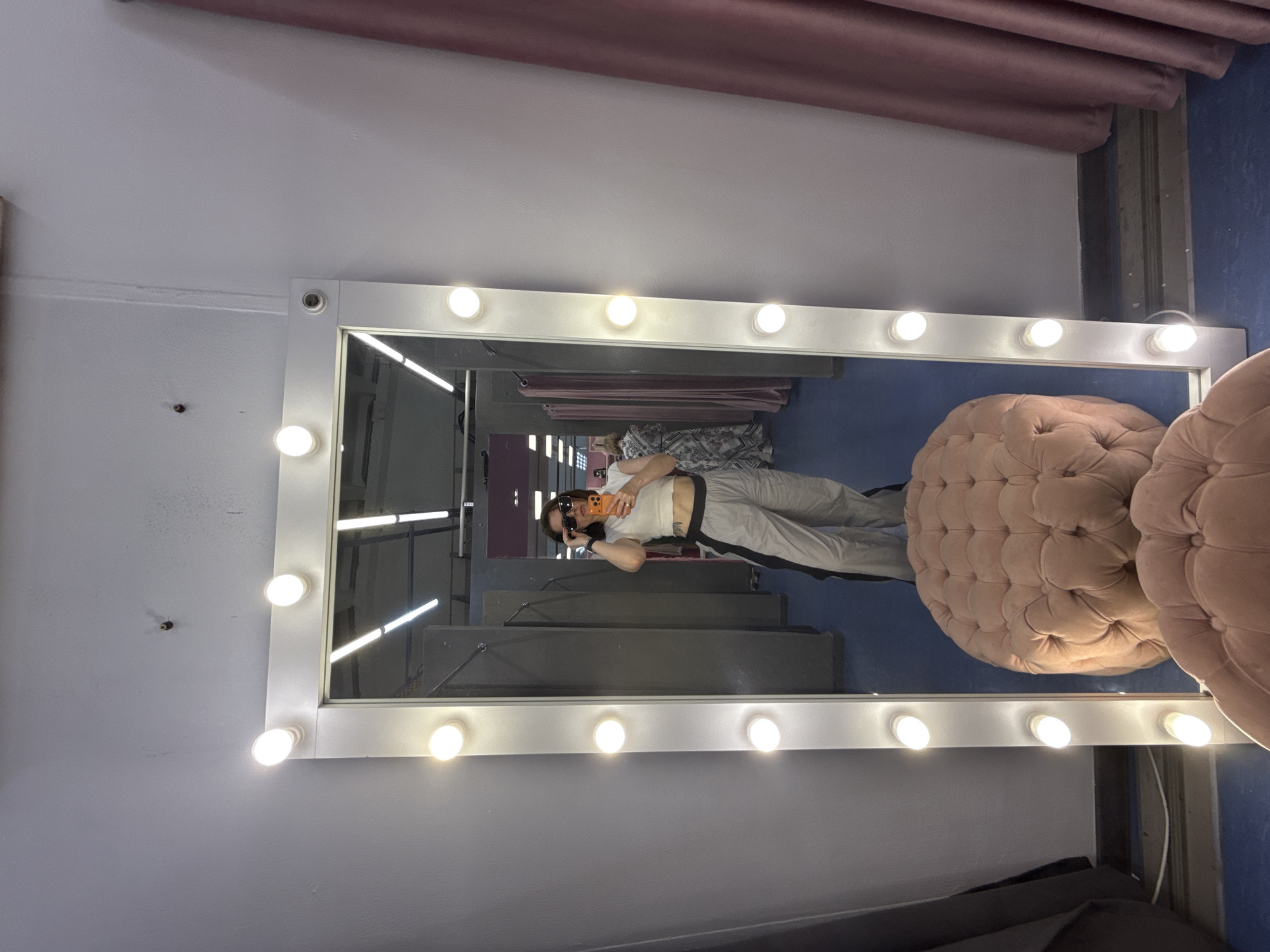 The image shows a reflection in a large, illuminated makeup mirror. The reflection features a person wearing sunglasses, a white shirt, and light-colored pants with a dark stripe, holding a mobile phone. The person appears to be taking a photo in the mirror.
Behind the person, in the mirror's reflection, there are some clothing racks and other upholstered furniture in the background. A pinkish upholstered pouf sits to the right of the mirror. The surroundings suggest the location is most likely in a fitting room or a clothing store. The location in Izhevsk, Russia cannot be confirmed without further data in the image.
