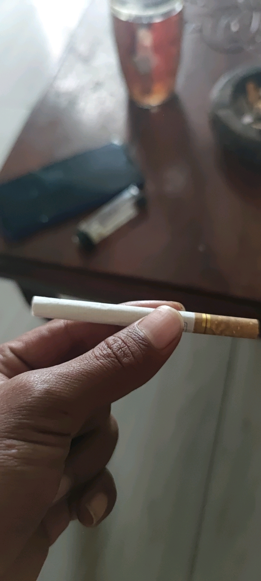 A person's hand holds a cigarette, with the tip of the cigarette featuring a gold band. In the background, on a wooden surface, a dark-colored smartphone and a lighter are visible. To the right, a glass filled with a dark amber liquid sits near an ashtray containing a lit cigarette. The scene is illuminated by ambient light, casting soft shadows. The location is specified as Gambarsari, Indonesia.