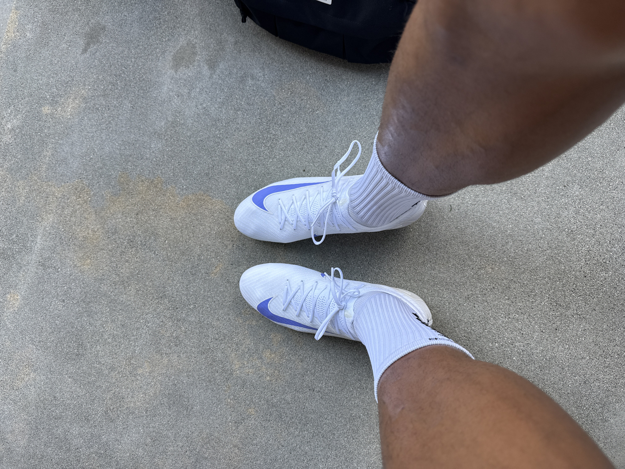 The image captures a close-up, overhead view of a person's lower legs and feet on a light grey, granular ground surface, likely concrete or asphalt. The person, who has a dark skin tone, is wearing white, ribbed athletic socks and a pair of white high-top athletic shoes, possibly cleats or trainers. Each shoe features a distinct purple Nike "swoosh" logo on its outer side and is secured with white laces. A portion of a black backpack is visible on the ground in the upper left quadrant of the frame, near the person's left leg. The lighting indicates it is daytime, with bright and even illumination. The person appears to be standing still. The setting is outdoors, possibly in an urban or sporting environment in Los Angeles, United States of America.