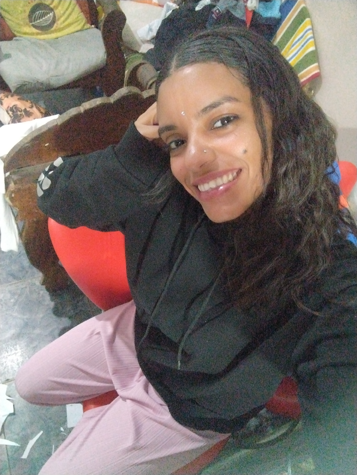 The photo shows a woman in a black hoodie and light pink pants seated on a red chair. She has long, dark, curly hair and is smiling. There is a small nose ring in her left nostril and a small bindi on her forehead. The background includes a wooden table, a pillow with a leaf pattern, and a variety of clothing items. The floor is tiled. The location is identified as El Valle del Espíritu Santo, Venezuela.