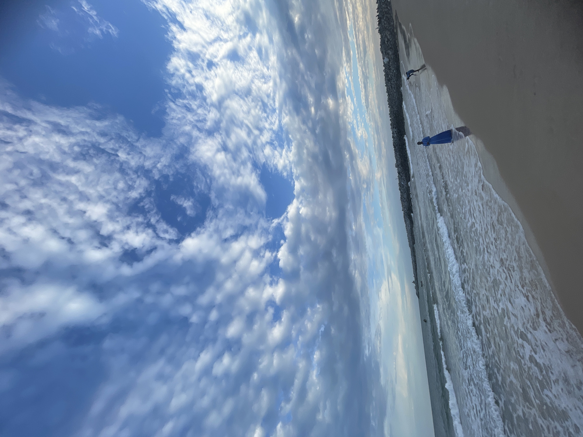 The image shows a beach in Ilado, Nigeria. The sky is blue with scattered white clouds, featuring a circular clear patch. The beach meets the ocean with gentle waves. Two people are visible: one figure further away and another person wearing a blue dress closer to the viewer. A rocky barrier runs along the edge of the beach.