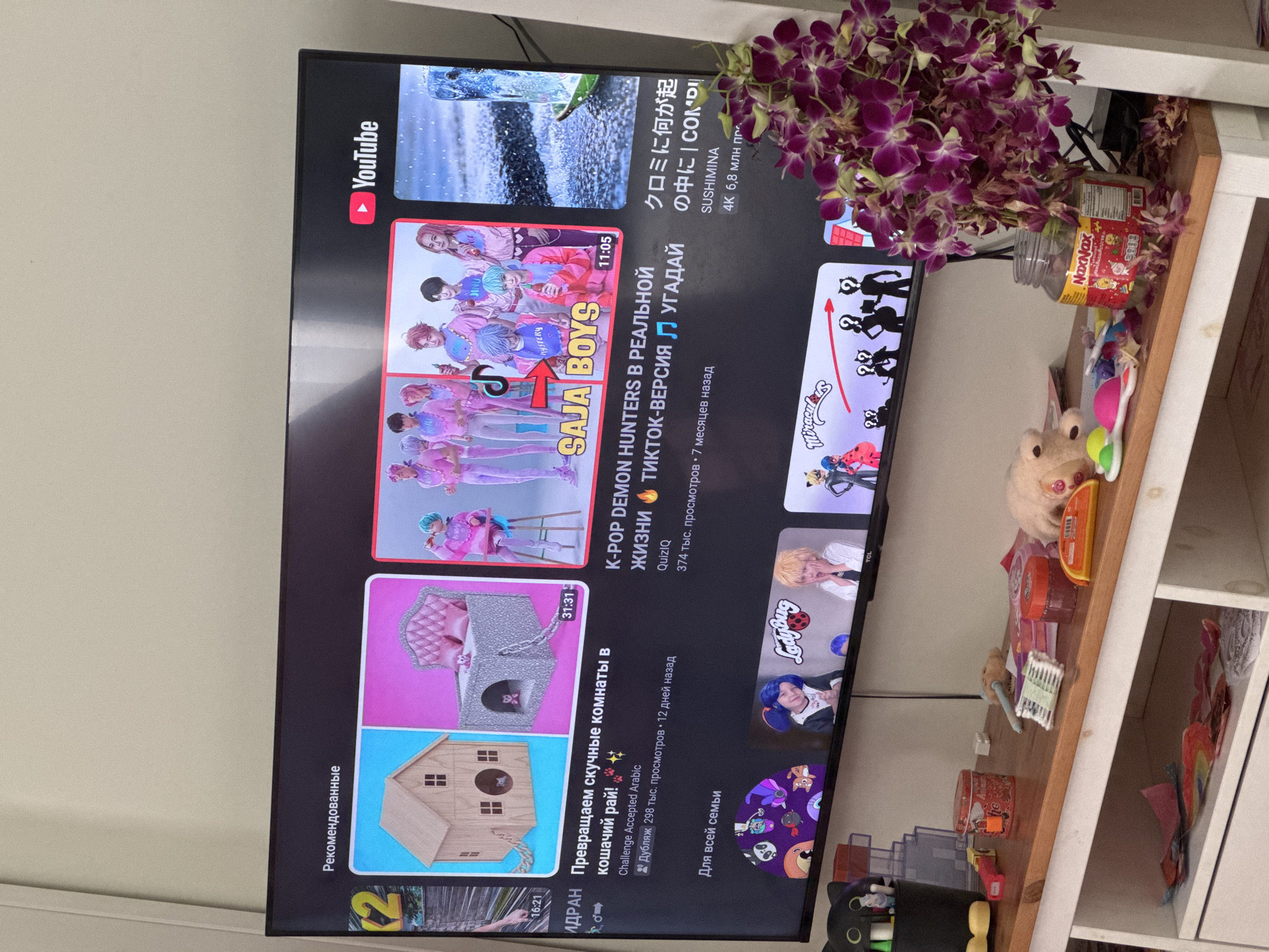 The image shows a YouTube interface on a TCL television screen, with recommended videos including a SAJA BOYS video and "Ladybug" content. The language used is primarily Russian and Japanese. To the right of the TV, there's a shelf containing items like a jar of "NaxNax," assorted toys, and a black cat-shaped pencil holder. There's also a bouquet of purple orchids placed near the TV. This scene is occurring at an unspecified location in Pa Khlok, Thailand.