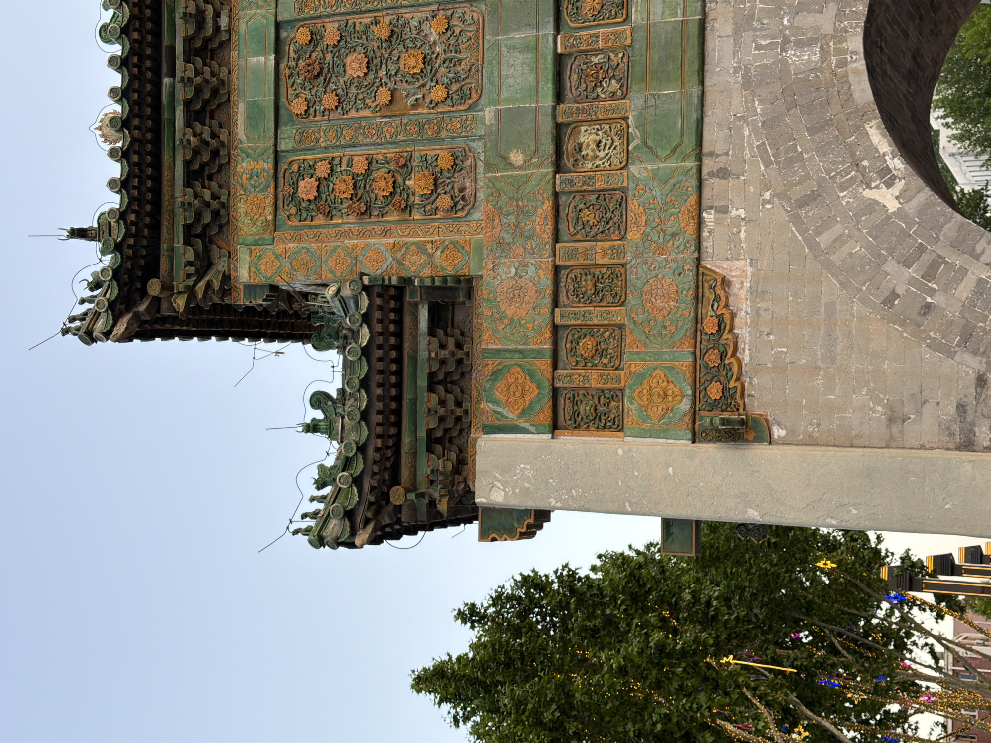 The image shows a close-up of an ornate structure with traditional Chinese architectural elements. The structure features green and gold detailing, particularly in the form of floral and geometric designs on tiled panels. There is a section of brickwork with an arched opening visible on the right side of the frame. A tree with green foliage, partially decorated with festive lights, is seen in the lower part of the image against a pale sky. This structure is located in Chaoyang District, China.