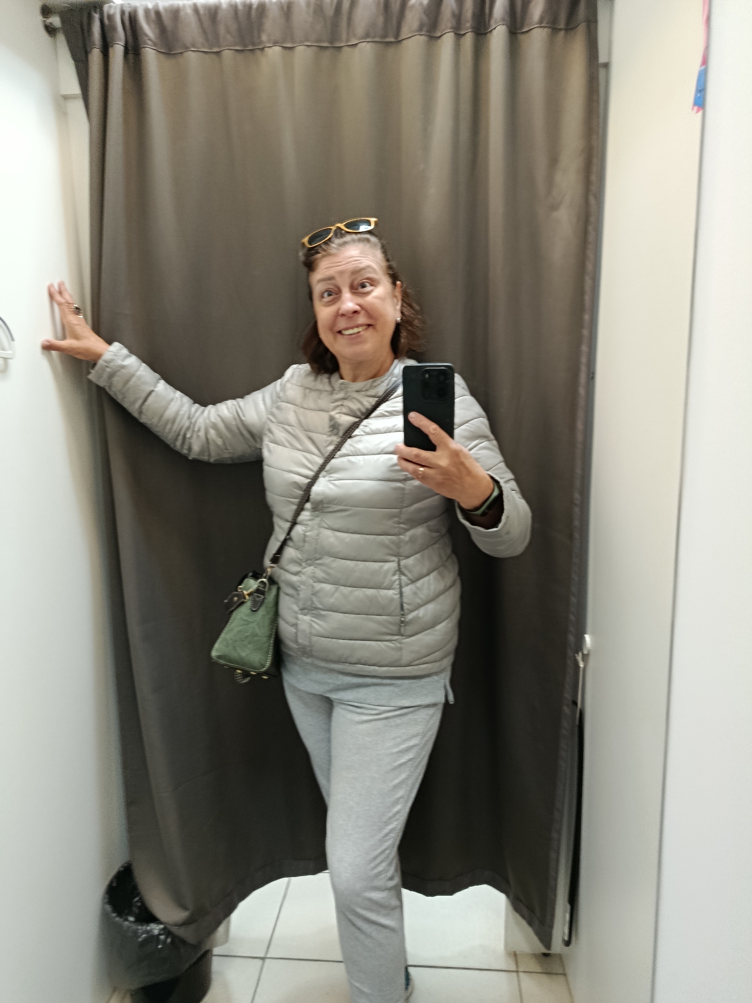 A woman with brown hair is standing in a fitting room, holding a smartphone to take a selfie. She is wearing a silver puffer jacket, grey sweatpants, and a green crossbody bag. Her sunglasses are perched on her head. She is smiling and looking at the camera. The fitting room has grey curtains and white walls. A trash can is visible to the left of the woman. The image does not contain any contextual information that would suggest a location in the city of Верея, Russia.