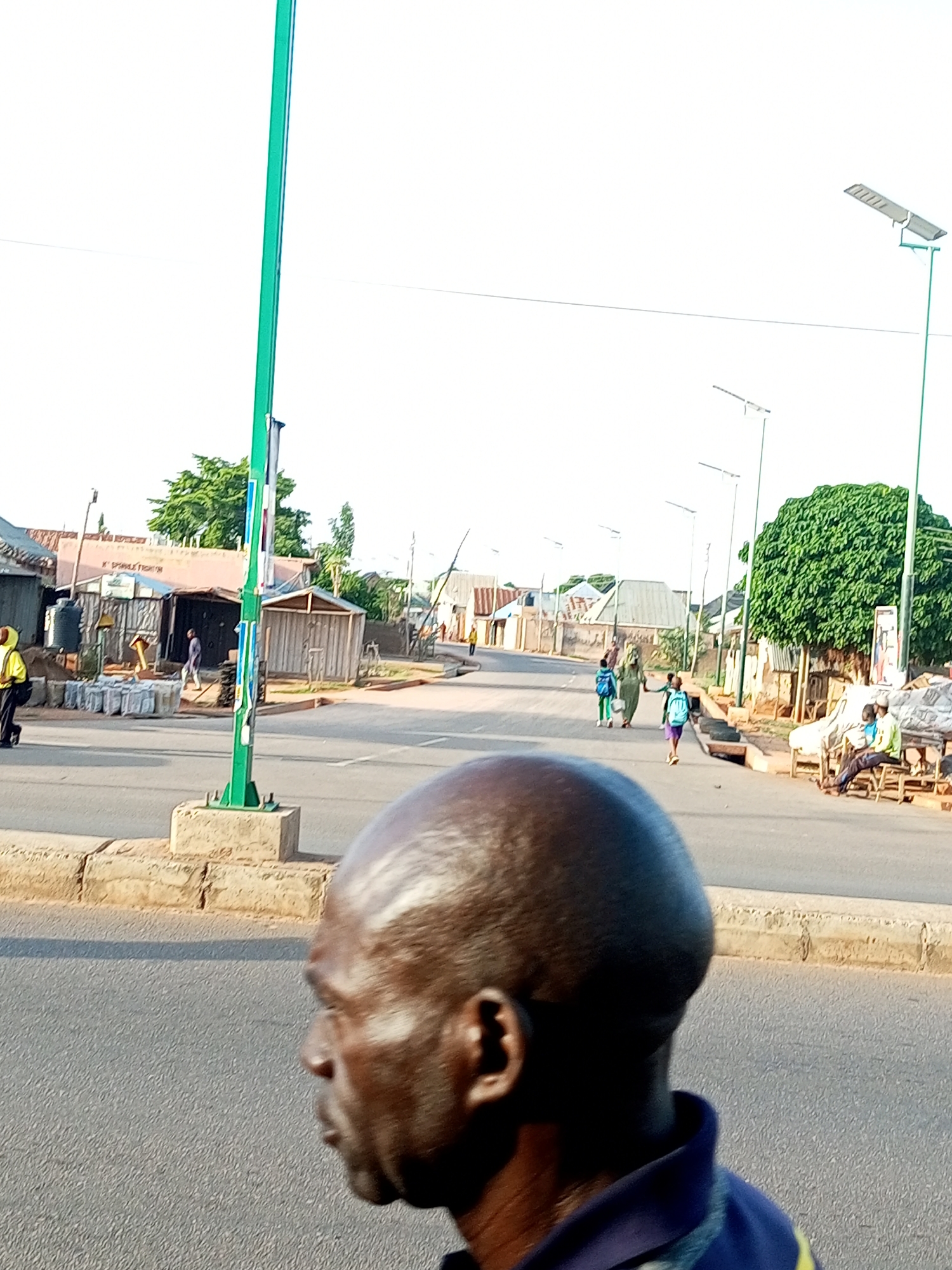 The image captures a street scene in Tumfure, Nigeria, during what appears to be daytime with a bright, overcast sky. In the foreground, a man's head and shoulders are visible in profile, facing to the left. His dark skin is glossy, suggesting sweat or oil, and his head is bald and shiny. He is wearing a dark blue polo shirt with a lighter blue collar and a yellow stripe on the sleeve.
The background depicts a street with several people walking. To the left, a person in a yellow top and black pants is walking away from the camera. Further down the street, there are shops and structures made of corrugated metal and wood, some with open fronts. A prominent green pole, possibly a lamppost or utility pole, stands near the center of the image, with several flyers or posters attached to it.
Further down the street, a group of children and adults are walking. One child wears a blue backpack, and another is dressed in a light-colored garment. A large, leafy green tree stands on the right side of the street, providing shade to some of the structures. In the distance, more buildings and trees are visible, and several modern streetlights with solar panels line the road. On the right side of the road, two men are seated on a bench outside a shop. One man wears a lime green shirt and is looking towards the street. The other man, in a blue shirt, is looking down.
The overall atmosphere suggests a normal day in a town or city. There are no overt signs of distress or unusual activity. The presence of modern streetlights indicates some level of development. No visible text is legible in the image.