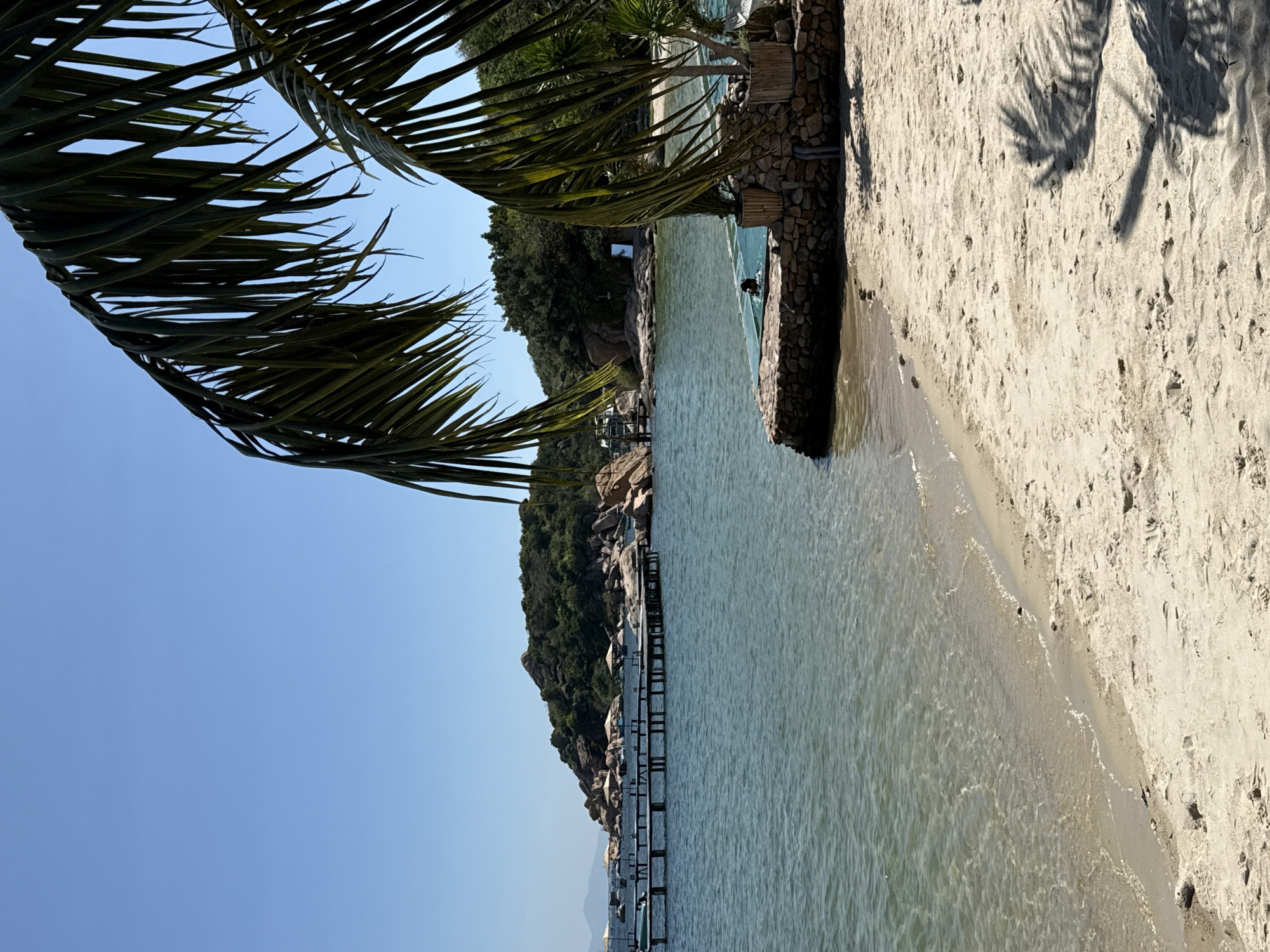 The image depicts a sunny coastal scene in Cam Ranh, Vietnam. A clear blue sky occupies the upper portion. The calm, turquoise water of a bay fills the left and central mid-ground, featuring gentle ripples. A light-colored sandy beach extends across the lower right and foreground, showing footprints and the distinct shadow of a palm frond.
In the mid-ground, a lush, green, rocky hill rises from the water on the left, with numerous large boulders along its base. Extending into the bay from the left is a wooden pier with multiple sections, some supporting white umbrellas. A small, light-colored boat is moored near the pier.
On the right side of the image, bordering the beach, a stone structure encloses a clear-sided pool or swimming area within the bay. A single individual is visible, partially submerged and swimming within this enclosure. Smaller potted plants are situated around the stone structure. Overhanging palm fronds frame the top and right sides of the image, obscuring parts of the sky and coastline. A rectangular, reflective object is partially visible on the distant right shore, amidst the vegetation.