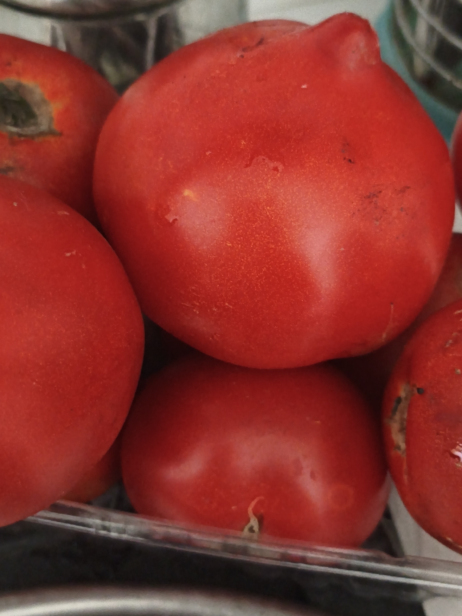A close-up, overhead shot captures a cluster of ripe, red tomatoes, some showing slight imperfections like dark spots and faint blemishes. The tomatoes are arranged in a clear plastic container, with some of them spilling over the edge, obscuring the bottom half of the container. The background is blurred, suggesting an indoor setting. The image's composition highlights the texture and vibrant color of the tomatoes, with one prominently featured in the center, filling most of the frame. The lighting casts subtle shadows, emphasizing the roundness of the fruits. There are no people or overt actions visible. The scene is consistent with a domestic kitchen or a market stall.
