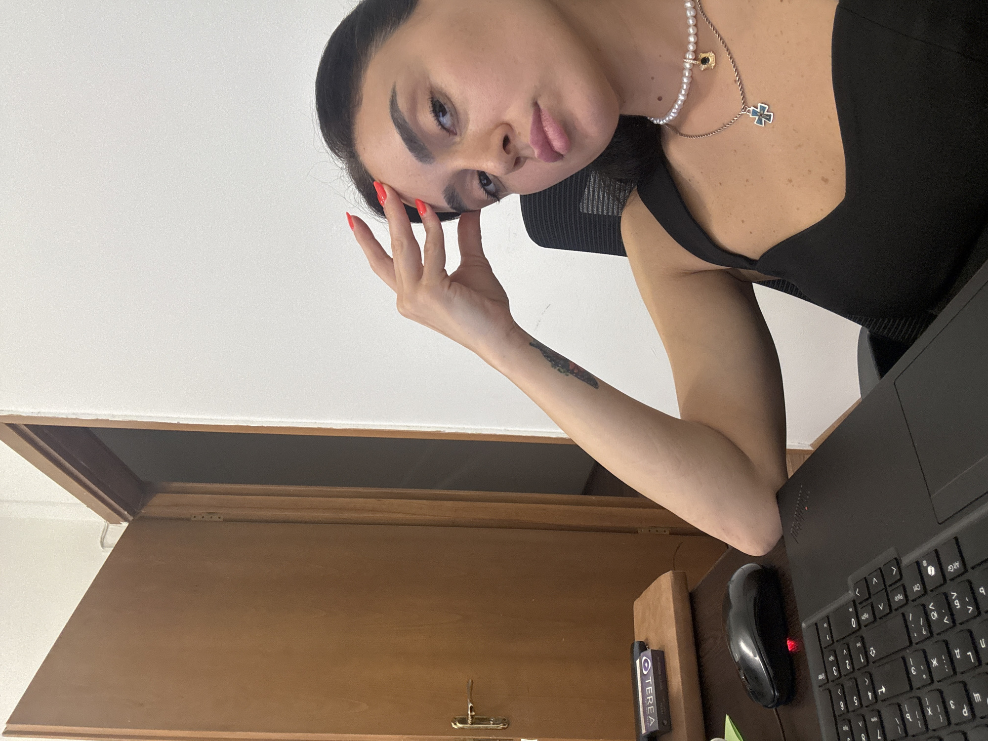 A woman is sitting at a desk in what appears to be an office or study. She is looking towards the camera with her hand on her forehead. A laptop, a computer mouse, and a small box are on the desk in front of her. A wooden door is visible in the background. The overall lighting suggests an indoor setting, possibly during the day. The image does not contain any information that would indicate the location is in Tbilisi, Georgia.