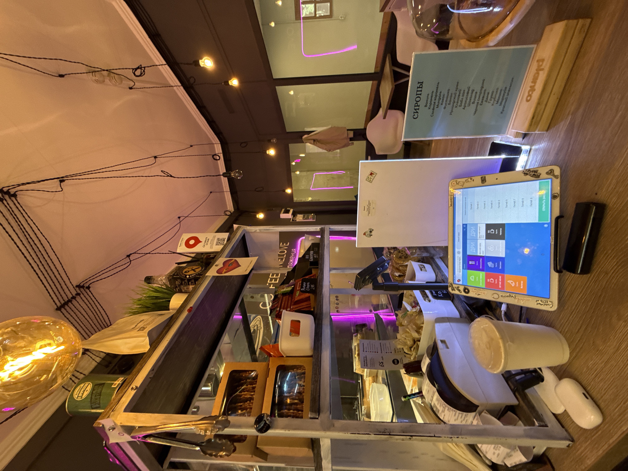 The image depicts the interior of a cafe in Moscow, Russia. In the foreground, a wooden counter displays a white tablet with a touch screen interface displaying a point-of-sale system, a disposable coffee cup with a lid, and a white charging case for wireless earbuds. To the right of the tablet, a light blue informational sign titled "СИРОПЫ" lists various syrup flavors in Russian. Behind the counter, a shelf unit contains various items, including snacks in boxes, a white container, a bottle of liquor, and a decorative plant. The cafe's ceiling is adorned with black electrical wires and several Edison-style light bulbs, with a large, glowing pendant light fixture prominently featured. Neon pink lighting accents the interior walls and windows. The overall ambiance suggests a modern, casual cafe environment.
