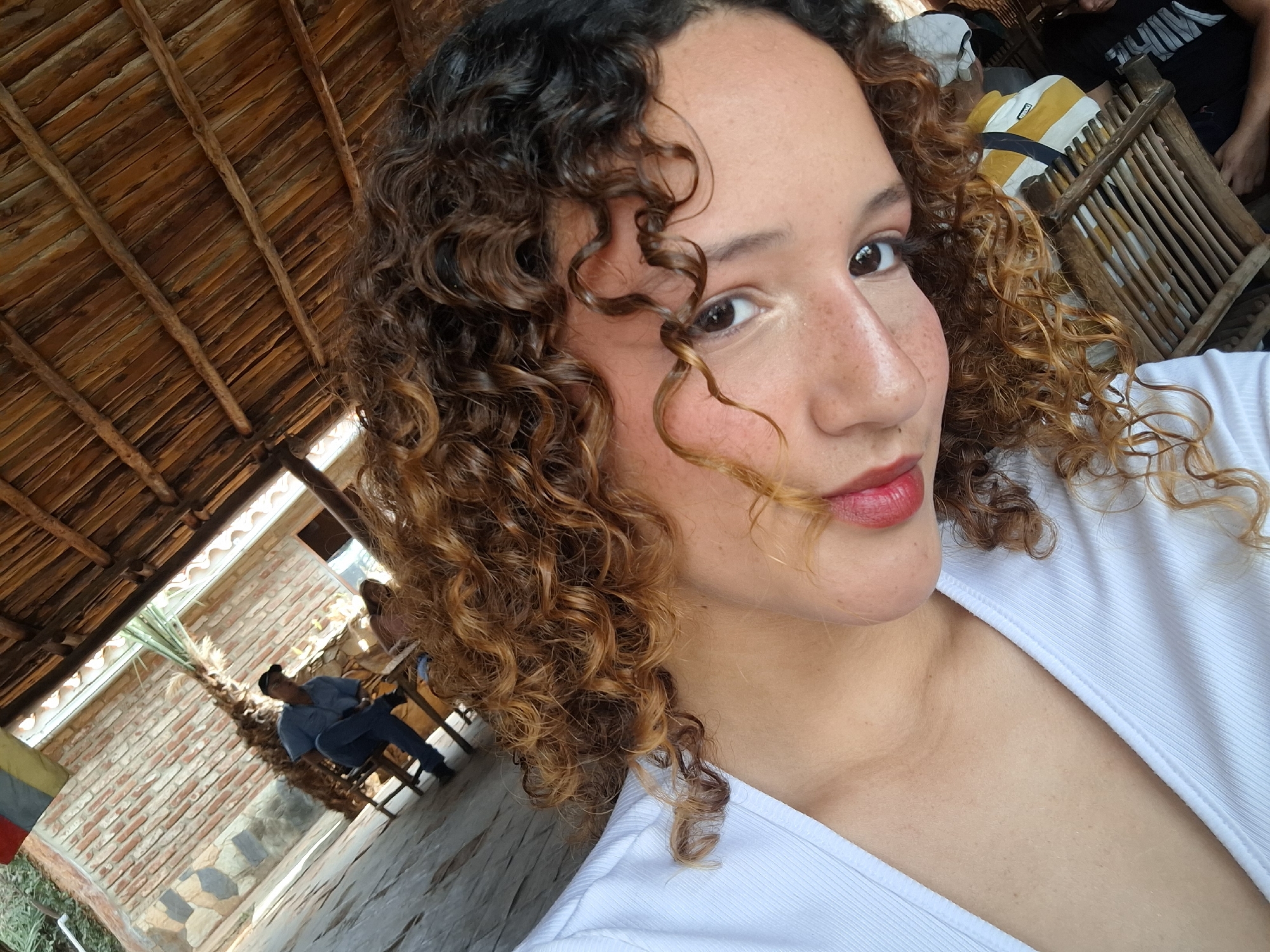 A close-up shot of a woman with curly brown and blonde hair, freckles, and red lipstick. She is wearing a white shirt. The background reveals a wooden roof structure and, through an opening, a man sitting outside in what appears to be a patio or outdoor dining area of a building constructed with brick. The location is Unknown, Unknown.