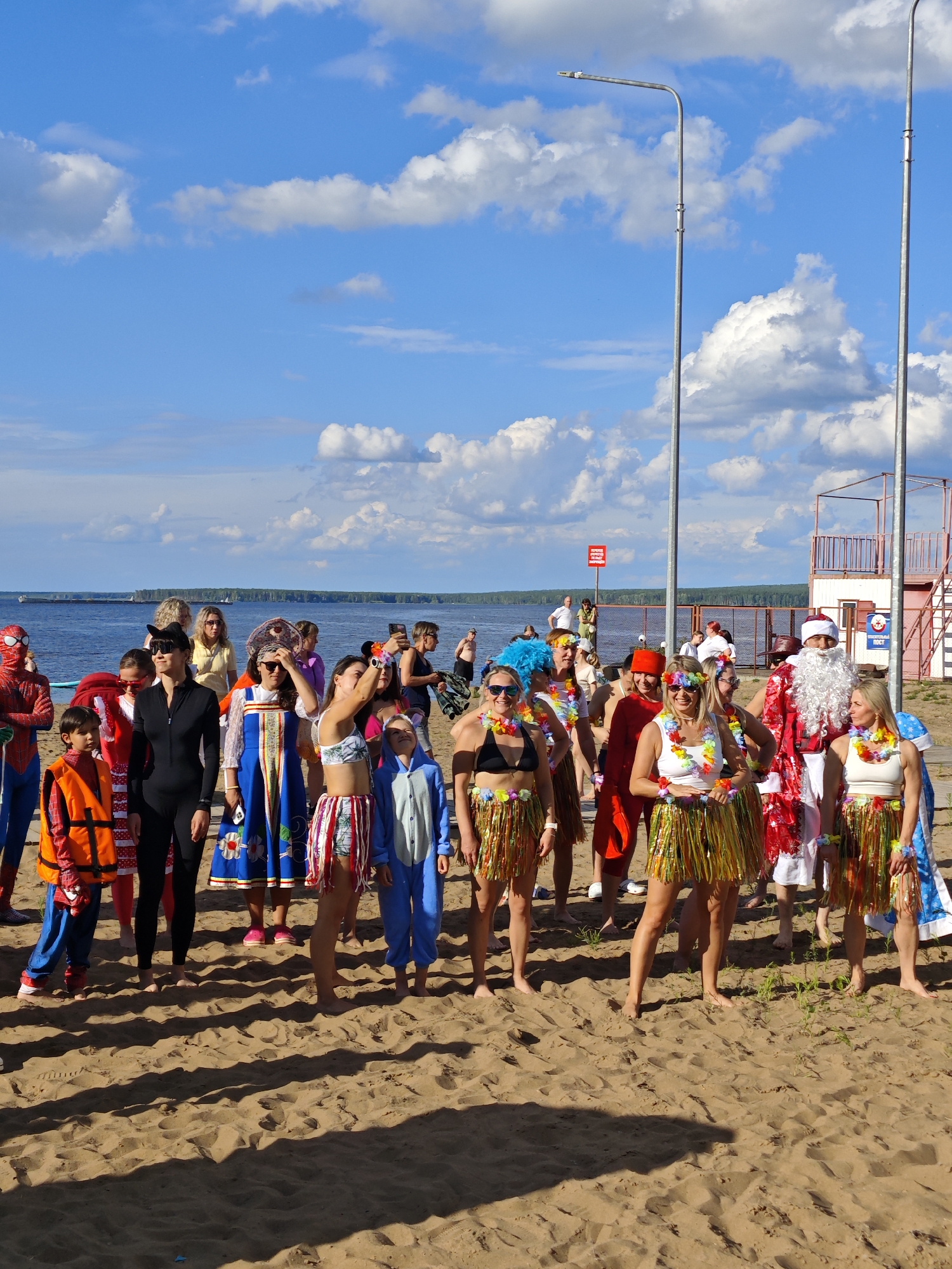 A group of people in costumes are gathered on a sandy beach in front of a body of water. The sky is blue with fluffy white clouds. Two tall lamp posts stand on the beach, and in the background, there is a red and white structure that appears to be a lifeguard tower or a small building with stairs. The people are dressed in a variety of outfits, including a Spider-Man costume, a black catsuit, a traditional blue and embroidered dress, a blue animal onesie, Hawaiian-style grass skirts with leis, and a Santa Claus costume. Some individuals are wearing sunglasses and leis, suggesting a festive or celebratory event. The lighting indicates it is daytime, likely late morning or afternoon, with bright sunlight casting shadows on the sand. The scene appears to be a public beach, possibly in Chaikovsky, Russia, given the context provided. The overall mood seems cheerful and casual, with people participating in a themed gathering or event on the beach. There is a red sign with white text visible on the fence behind the group of people, but the text is not legible.