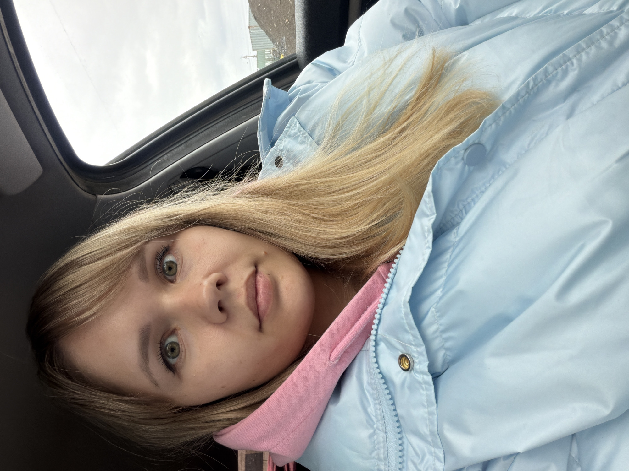 A young woman with light blonde hair and green eyes is prominently featured in a close-up shot, angled diagonally. Her gaze is directed towards the camera with a wide-eyed, somewhat surprised or curious expression. Her lips are slightly parted. She has fair skin and visible eyelashes, suggesting light makeup.
She is wearing a light blue puffer jacket over a pink hoodie, with the pink fabric visible around her neck and chest area. Her long blonde hair flows across her shoulder and chest.
The setting appears to be inside a vehicle. Above her, a car's sunroof or panoramic roof is visible, showing an overcast, cloudy sky. To her right (viewer's left), a car window and its frame are present, through which a blurred outdoor background, possibly with some distant structures or a rough ground, can be seen. The overall image has a tilted orientation, suggesting the photo was taken from a low angle or while reclining. No text is visible in the image.