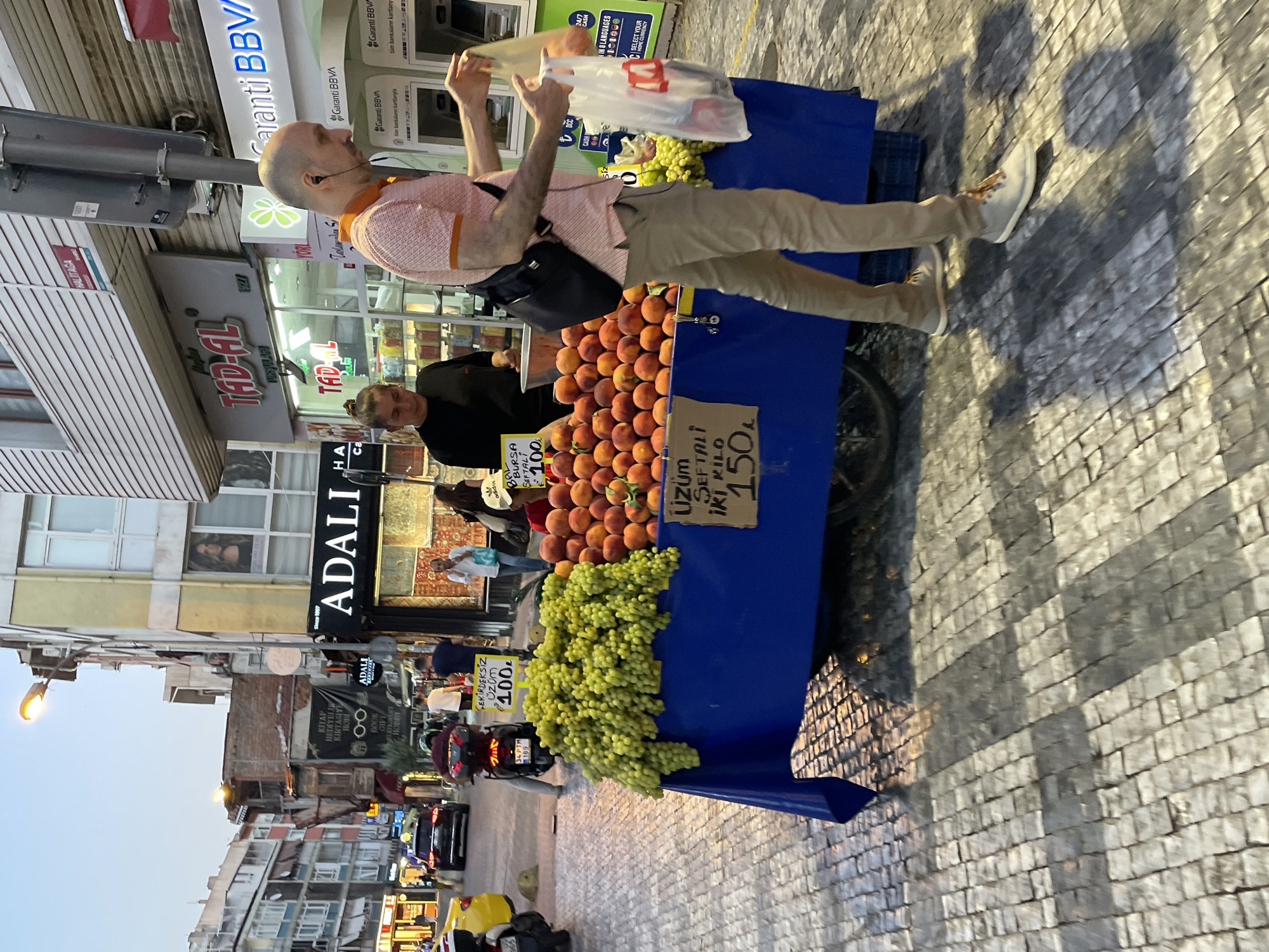 In Kadıköy, Turkey, a street scene unfolds with a fruit stand as the central focus. A man, wearing a pink patterned polo shirt and beige pants, is captured mid-action, reaching into a white plastic bag he is holding. His bald head and dark earbuds are notable features. He stands to the right of a fruit cart laden with peaches and grapes. A woman with her hair in a bun, dressed in a black top, stands behind the fruit stand, appearing to interact with customers or manage the stall. The fruit stand itself is draped with a bright blue cloth. Signs on the stand display prices, with one indicating "üzüm seftali kilo 150" (grape peach kilo 150) and another "üzüm 100" (grape 100). In the background, the street is paved with cobblestones, and buildings line the street, some with awnings and storefronts. A scooter with a license plate that reads "34 PTM 189" is parked to the left of the fruit stand. A yellow vehicle is visible further down the street, and traffic can be observed. The lighting suggests it is either early morning or late evening. The overall environment depicts a bustling urban market setting.