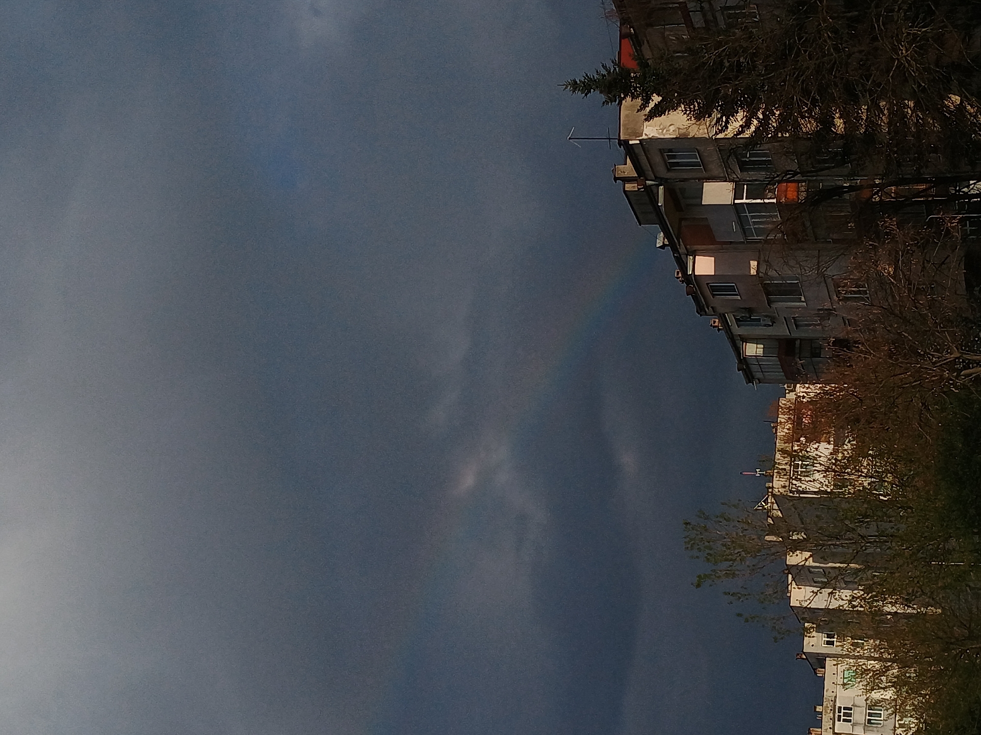 The image presents a vertical view of a cloudy sky with a faint rainbow arching in the center. The right side of the image features a series of residential buildings, possibly apartment blocks, which are characteristic of urban architecture in Veliko Tarnovo, Bulgaria. The buildings appear to be old, with multiple stories and windows, some with lights on, indicating occupancy. There are also trees with bare branches, suggesting the season may be autumn or winter. The overall scene evokes a sense of urban living under a dramatic sky.