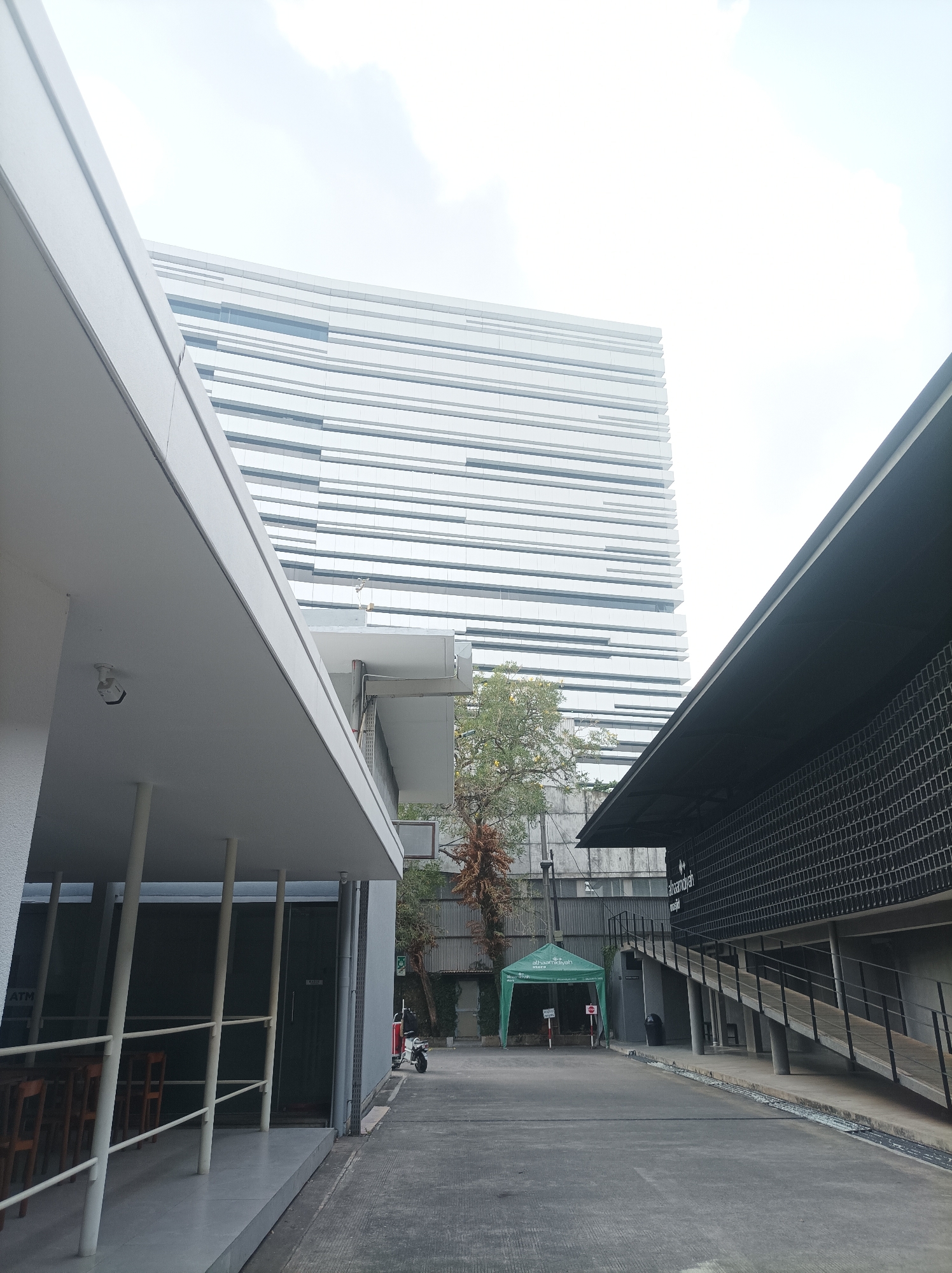 The image depicts a modern architectural scene in South Jakarta, Indonesia. In the foreground, a white, cantilevered structure with a sloping roof extends from the left side of the frame, providing shelter over a walkway with white railings and supports. Beyond this, a pathway leads into the midground where a green tent, possibly a temporary structure, is set up. Behind the tent, trees with visible dry foliage are present. Dominating the background is a tall, contemporary building characterized by its facade of horizontally layered white panels, creating a textured, striped appearance. To the right of the pathway, a dark, angular building features a patterned facade and an elevated ramp with railings, leading upwards. The sky is overcast, suggesting a diffused light. There are no people clearly visible in the scene. Objects include tables and chairs under the white structure, a motorcycle parked near the tent, and trash receptacles. The environment appears to be an outdoor urban space, likely a commercial or public area given the architectural styles and presence of a tent. The overall impression is one of clean lines and modern design.