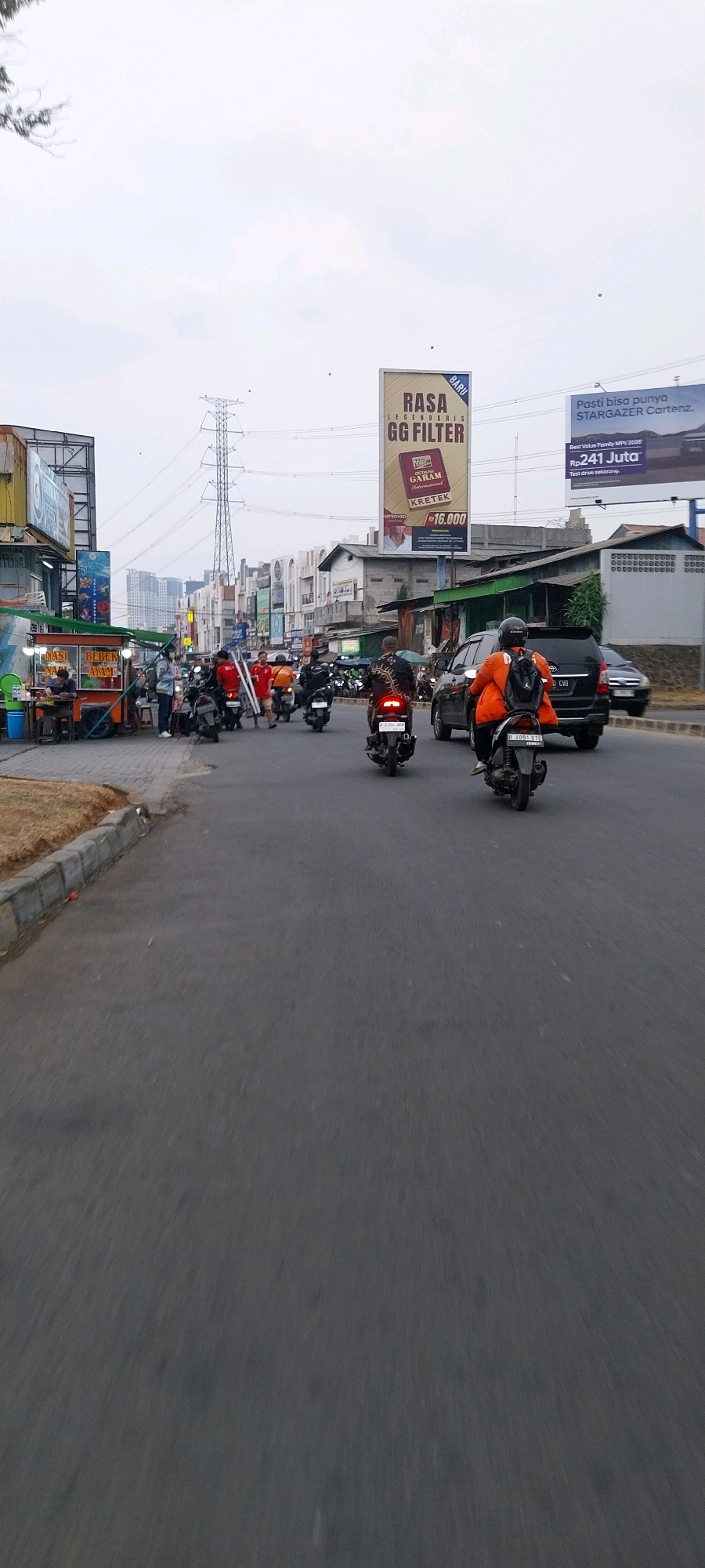 This is a street-level, eye-level photograph taken in Kelapa Dua, Indonesia, depicting traffic and commercial activity. The scene is dominated by a paved road with a slight incline, blurred due to motion. On the left side of the road, there is a sidewalk with a few food stalls, one of which displays "NASI BEBEK AYAM" (Duck and Chicken Rice). Several people are visible around the stalls, some standing and others seated.
Further down the road, a line of motorcycles and a few cars are in motion, suggesting moderate traffic. Two motorcyclists in orange jackets are prominent, one in the foreground and another further ahead. One motorcycle in the foreground has the license plate "B 6091 BTZ". A black SUV is also visible in the traffic.
Two large billboards are positioned above the buildings. The left billboard is partially obscured but appears to advertise something with "PT." and text. The larger right billboard prominently displays "RASA GG FILTER" in large letters, with smaller text below indicating "LEGENDARY" and "KRETEK" with a price of "Rp16.000". Below this, a portrait of a man is shown. To the right of this billboard, another billboard advertises "PASTI BISA PUNYA STARGAZER CARTEZ," referencing a car model and a price of "Rp. 241 Juta."
The environment suggests an urban or semi-urban setting with a mix of commercial establishments and residential buildings lining the street. Overhead, power lines and a tall transmission tower are visible against an overcast sky. The overall impression is a bustling daytime scene on a typical Indonesian road.