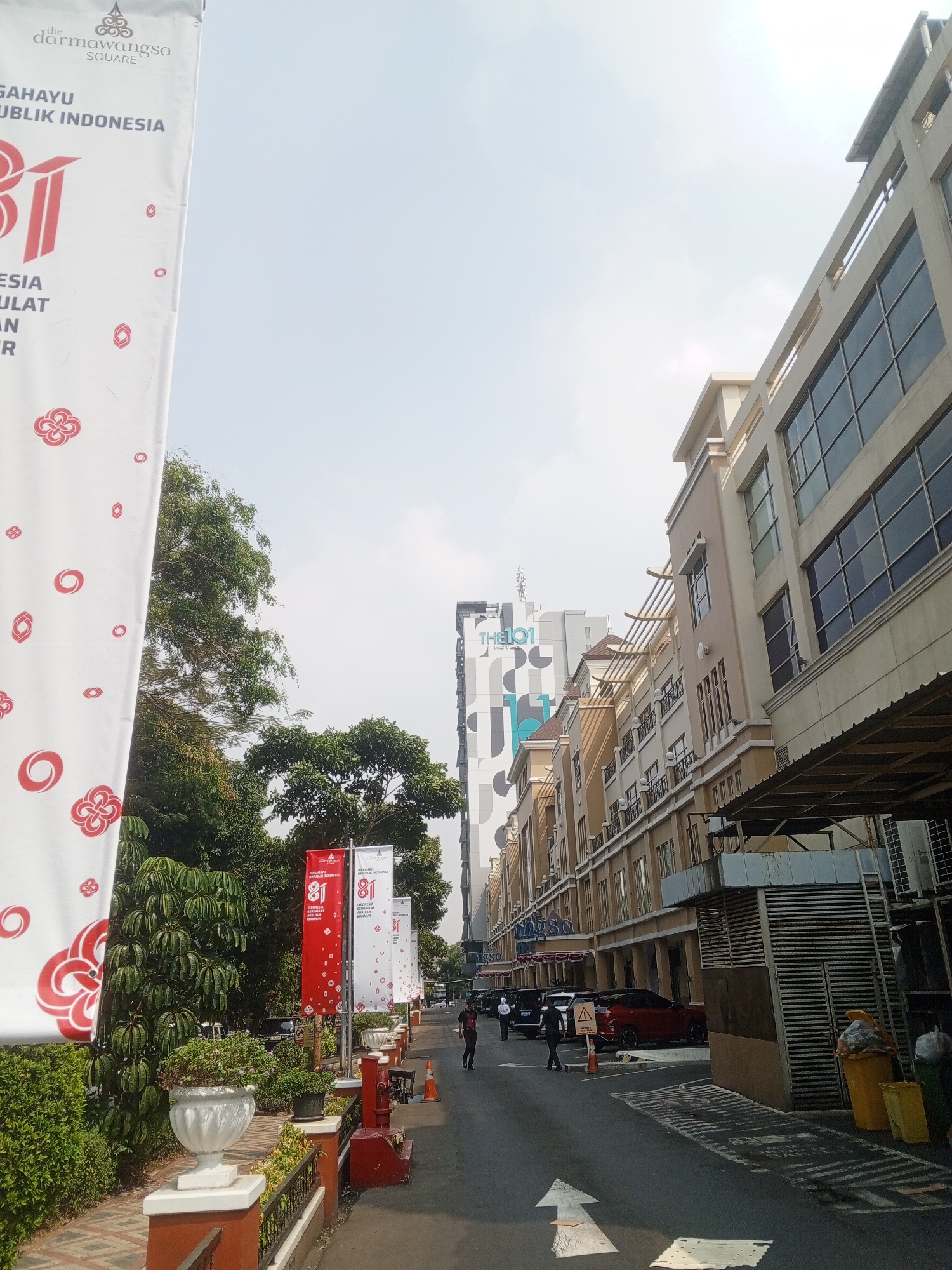 The image shows a street scene in South Jakarta, Indonesia, likely near the Darmawangsa Square. A white banner on the left side displays "THE DARMAWANGSA SQUARE" and "HUT RI KE-77" (77th Indonesian Independence Day) in Indonesian text and graphics. Red and white banners with "81" are displayed along the sidewalk. The buildings on the right appear to be commercial establishments, possibly shops or offices, with multiple stories and large windows. Several cars are parked along the street. A few individuals are walking on the sidewalk and in the street. The sky is clear and bright.